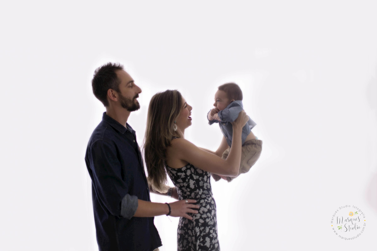 Fotografia de sessão de acompanhamento de bebê de 6 meses, com a mamãe o papai e o bebê no colo, eles estão de perfil, em fundo fotográfico infinito branco, todos estão sorrindo no studio localizado em Pinheiros, São Paulo, SP.