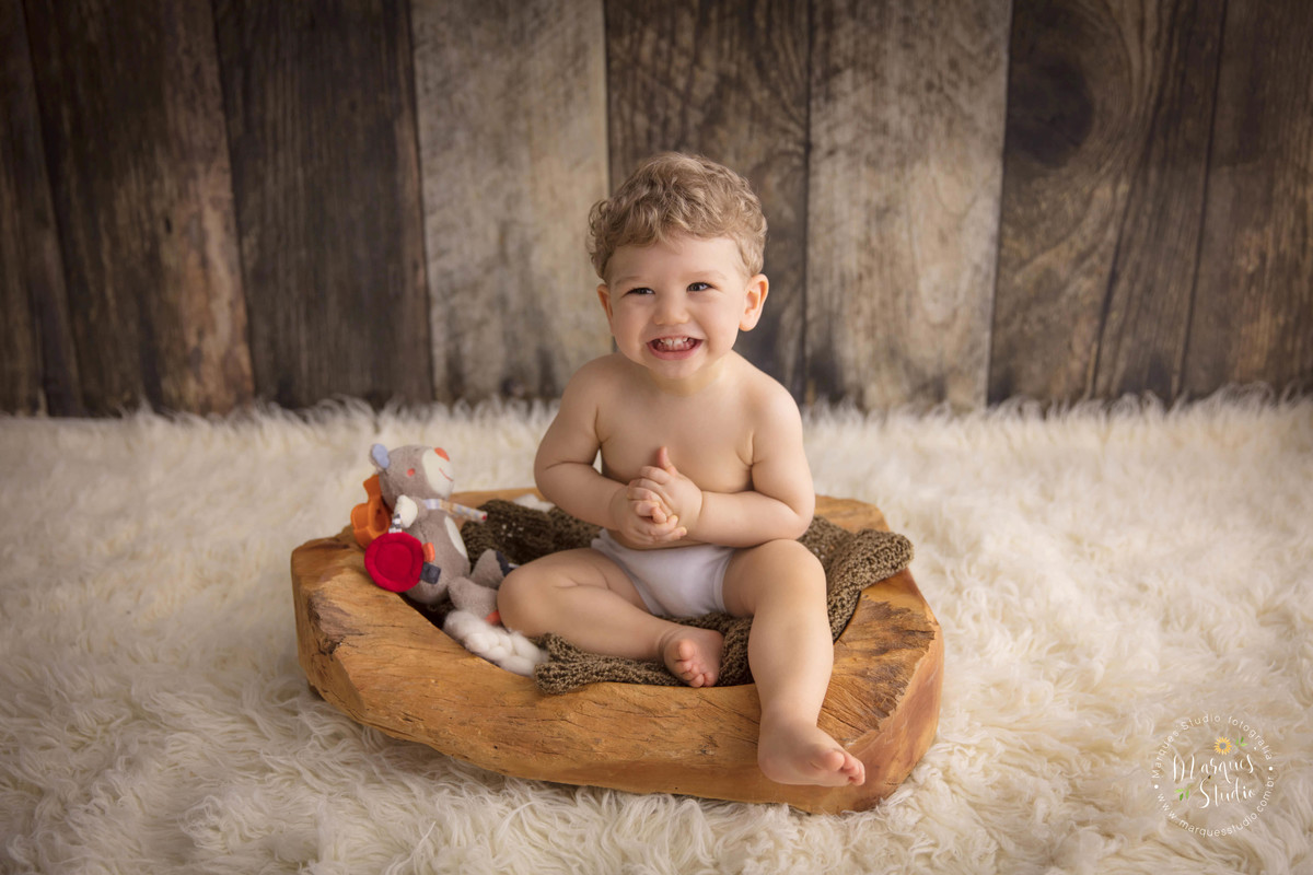 Foto feita em studio fotográfico localizado na zona Oeste de São Paulo, SP, com uma criança de 1 ano sentada em um cesto, o bebê está sorrindo, ele está batendo palma, e está em um lindo fundo fotográfico de madeira, com um lindo tapete bege.