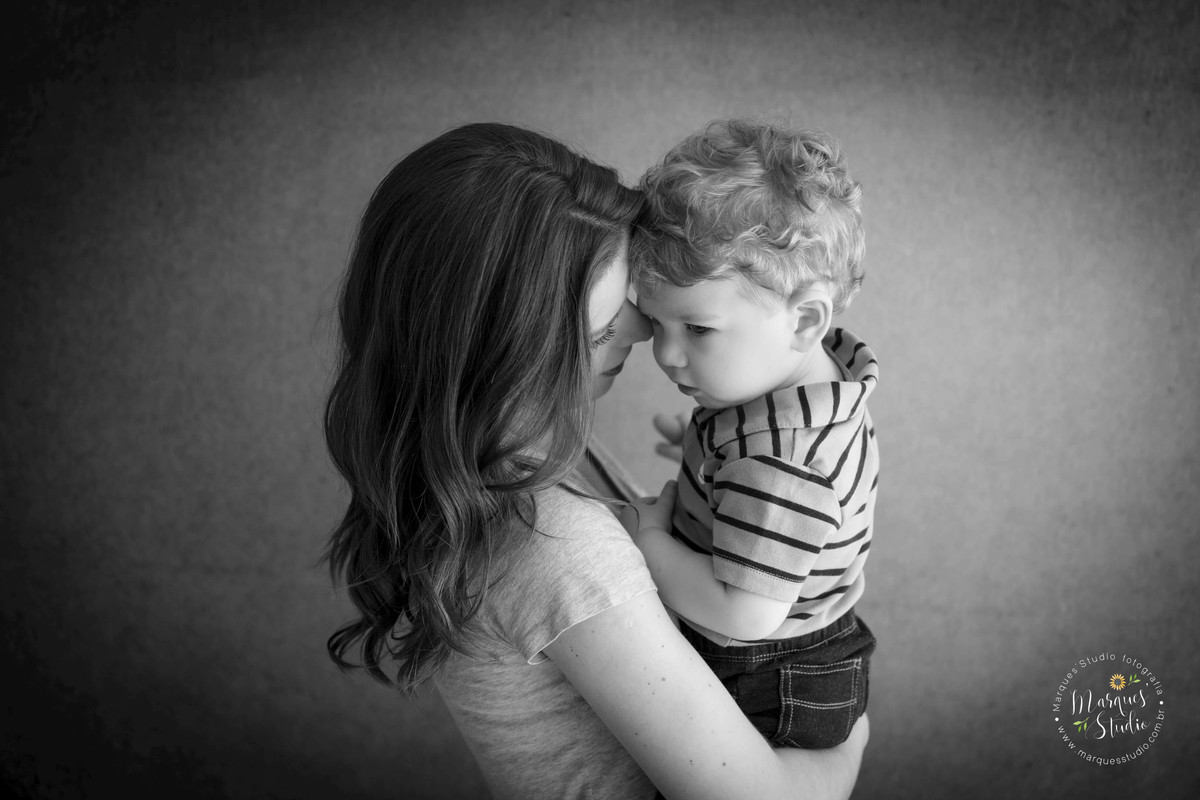 Foto da sessão de acompanhamento de 1 ano do Leonardo, no Studio, na Vila Madalena, São Paulo, SP, na foto temos a mamãe de perfil segurando o bebê no colo que está dando um lindo sorriso, eles estão no fundo fotográfico infinito preto.