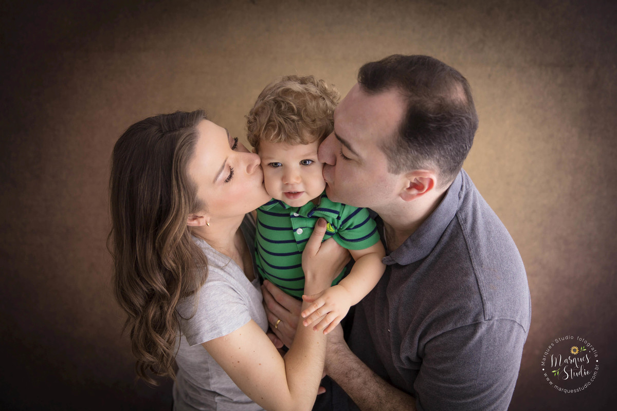 Fotografia da sessão de acompanhamento de 1 ano do Leonardo, no studio na Vila Leopoldina, São Paulo, SP, na foto temos a mamãe e o papai dando um abraço e um beijo na bochecha do bebê Leonardo, eles estão em um lindo fundo fotográfico bege.