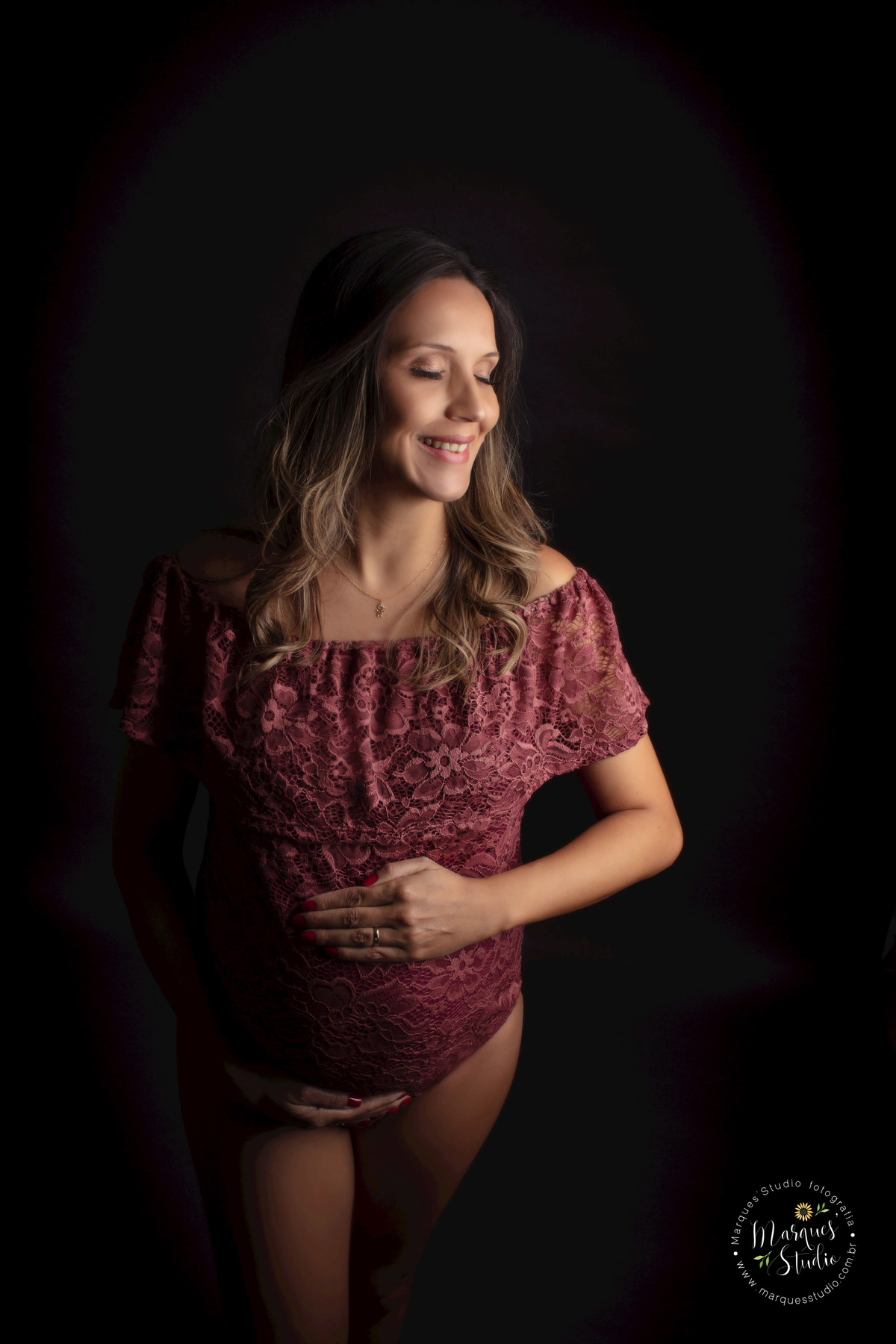 Foto feita em Studio na Vila Sumaré, São Paulo, SP, a gestante está em pé de perfil, ela está com as mãos sobre a sua barriga gravida com um belo sorriso estampado no rosto, ela está usando um body na cor marsala com renda, no fundo infinito preto.