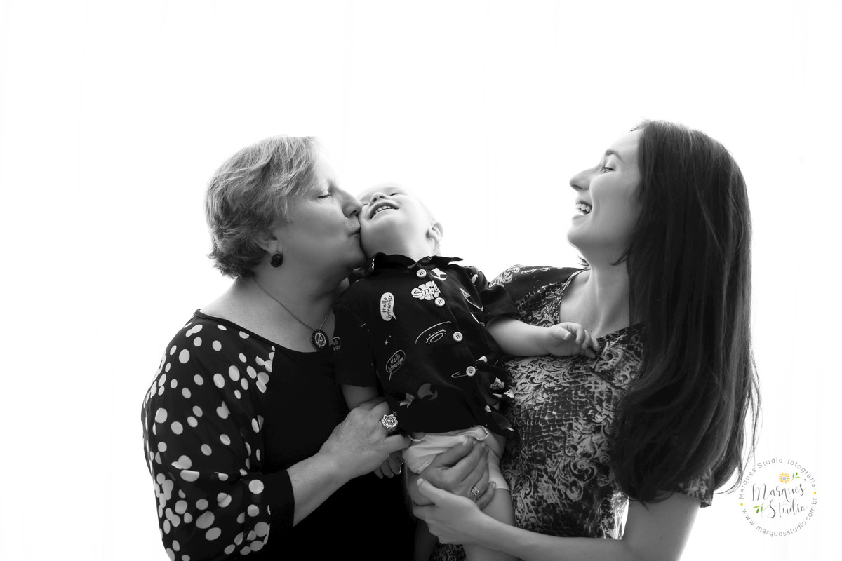 Fotografia da sessão de natal do Benjamin, no studio na Vila Leopoldina, São Paulo, SP, na foto temos a mamãe e a vovó do bebê dando um abraço e um beijo na bochecha do bebê Benjamin, eles estão em um lindo fundo infinito branco.