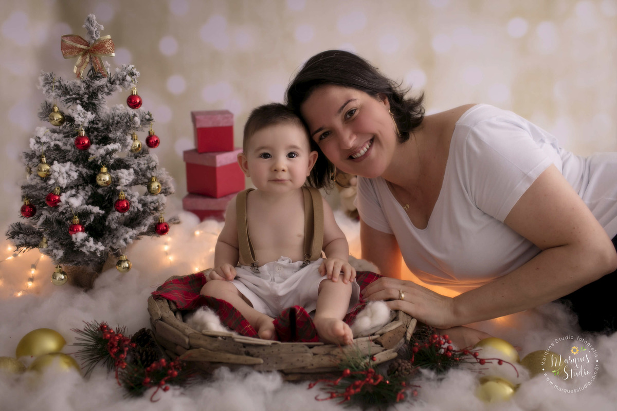 Fotografia de um ensaio de natal feita em estúdio fotográfico localizado na Av. Paulista, SP, na foto temos um lindo bebê de 6 meses sentado em um cesto, ao lado dele está sua mamãe, eles estão sorrindo, ao fundo temos uma arvore e luzinhas de natal.