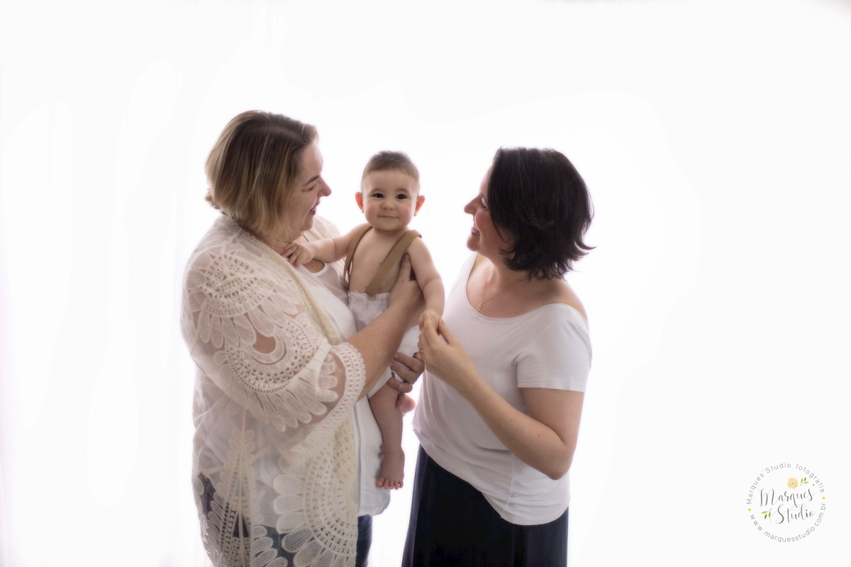 Fotografia da sessão de natal do Emanuel, no studio na Vila Leopoldina, São Paulo, SP, na foto temos a mamãe e a vovó do bebê dando um abraço olhando e dando um belo sorriso para o bebê Emanuel, eles estão em um lindo fundo infinito branco.