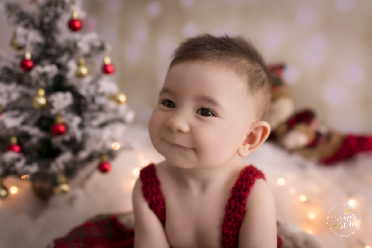 Foto Feita para a sessão de natal infantil em studio na zona Oeste, Sumaré, São Paulo, SP, de um bebê de 6 meses, ele está sentado em um lindo cesto, ao fundo temos uma linda arvore de natal, um papai Noel e luzinhas de natal ele usa um conjunto vermelho.