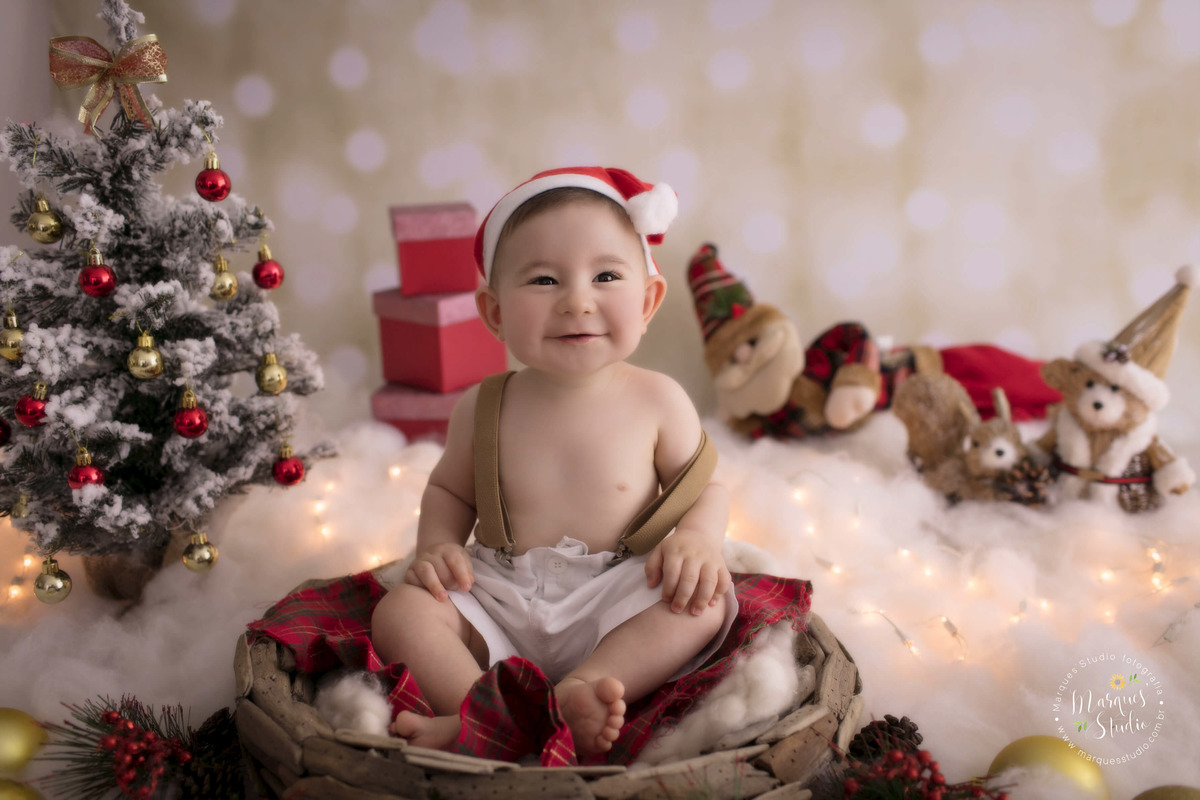 Foto feita no studio fotográfico em Osasco, SP, para o ensaio de natal do Emanuel, na foto ele está sentado em um cesto em um belo cenário com papai Noel, arvore e luzinhas de natal, ele usa um conjunto com suspensório bege e um lindo gorro de papai Noel.