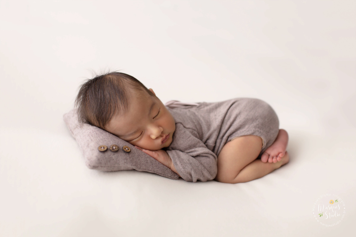 Fotografia de bebê recém-nascido menino, deitado no puff com as mãozinhas no rosto, foto feita em studio fotográfico na Vila Perdizes, São Paulo, SP, ele está dormindo e usando uma linda roupinha marrom, com o rosto em um lindo travesseiro marrom.