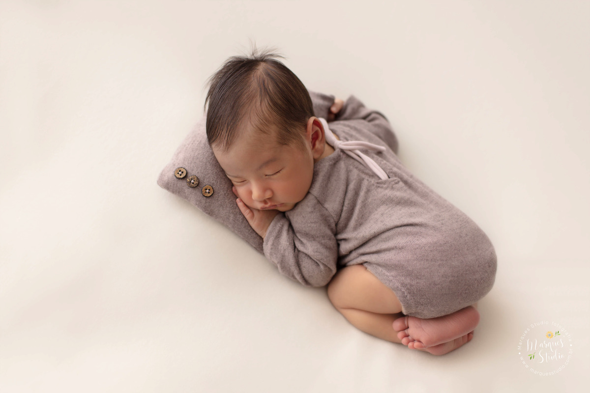Fotografia de bebê recém-nascido menino, deitado no puff com as mãozinhas no rosto, foto feita em studio fotográfico na Vila Leopoldina, São Paulo, SP, ele está dormindo e usando uma linda roupinha marrom, com o rosto em um lindo travesseiro marrom.