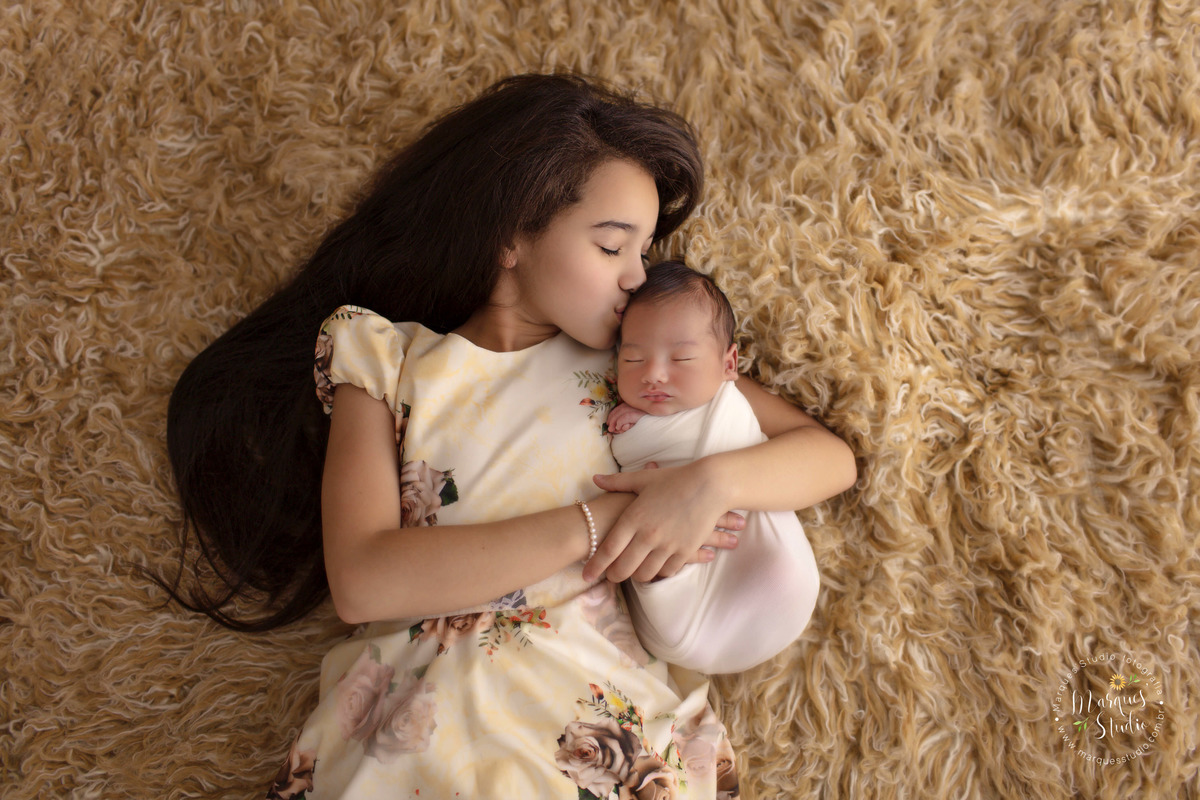 Foto de ensaio newborn,  recém-nascido abraçado com a irmã em ensaio fotográfico de bebê na vila Madalena em São Paulo, SP. Os irmãos usam roupinhas na cor bege, e estão deitados em um lindo tapete bege, a irmã está dando um beijo na cabeça do irmão.