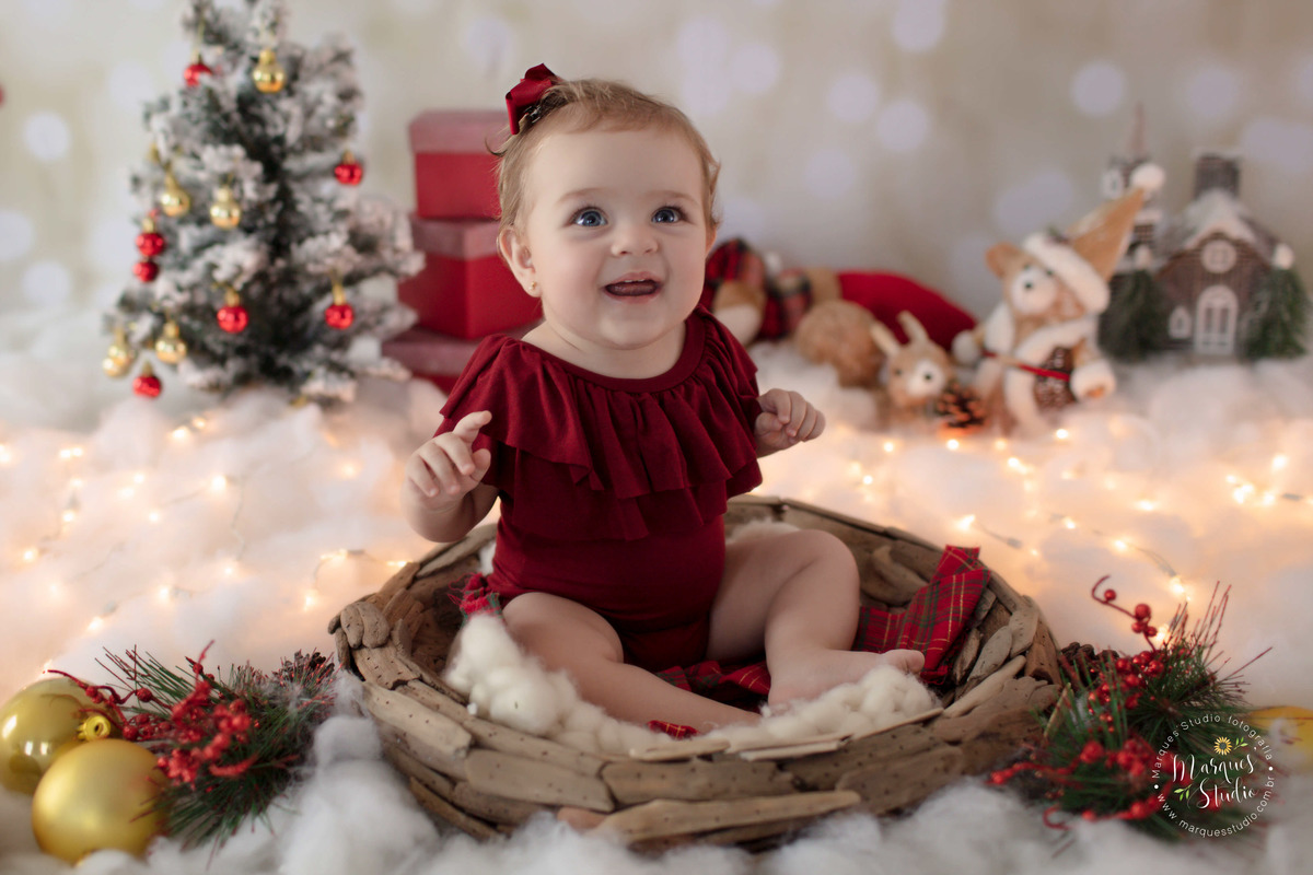 Foto feita no studio fotográfico, na Vila Leopoldina São Paulo, SP, para o ensaio de natal da Olivia, na foto ela está em um belo cenário com papai Noel, arvore e luzinhas de natal, com caixinhas de presentes, ela está usando um body vermelho, e sorrindo.