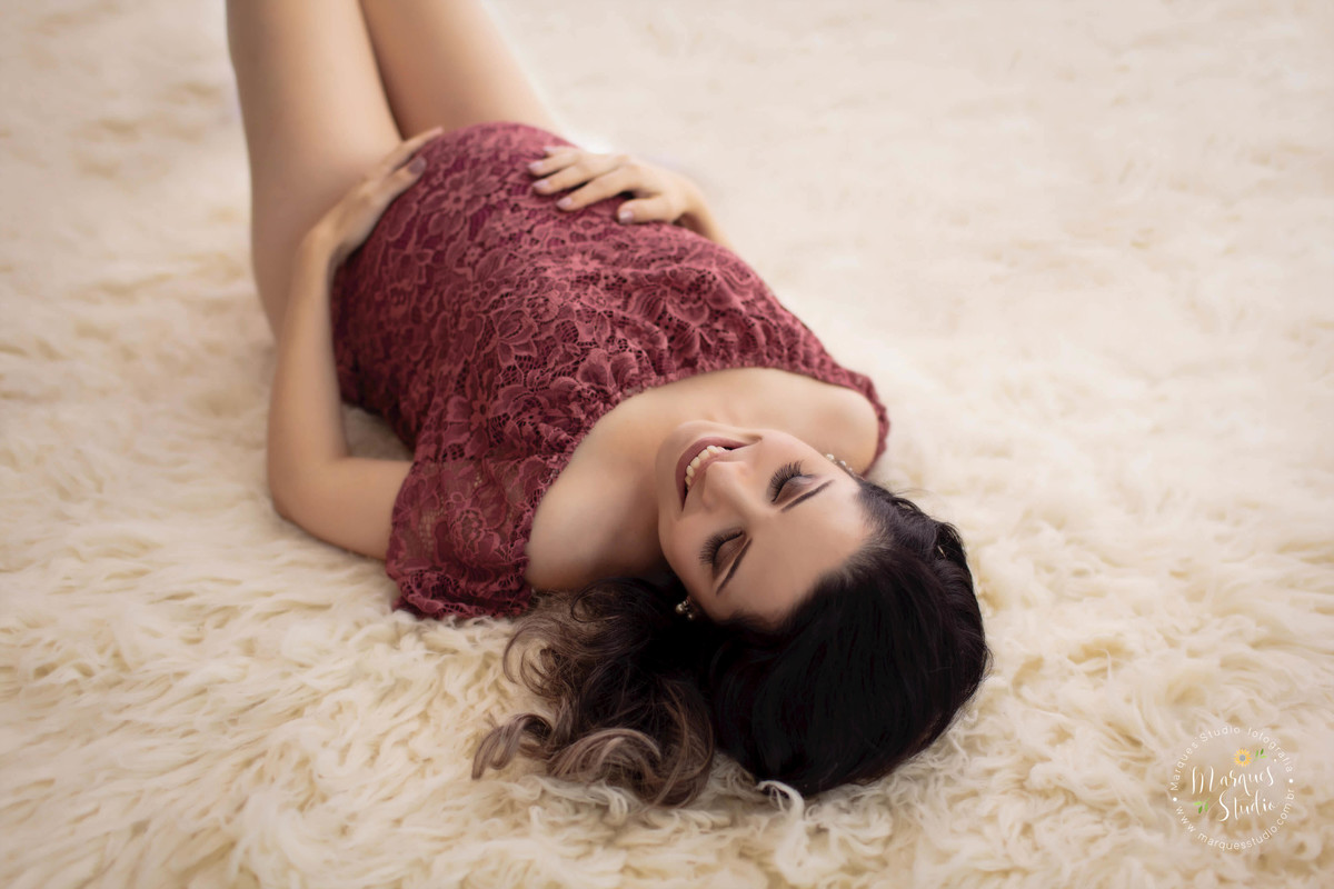 Fotografia de gestante em Studio na Vila Madalena São Paulo SP a gestante está deitada sobre um lindo tapete na cor bege ela está com as mãos sobre a sua barriga gravida com um belo sorriso estampado no rosto, ela está usando um body na cor marsala.