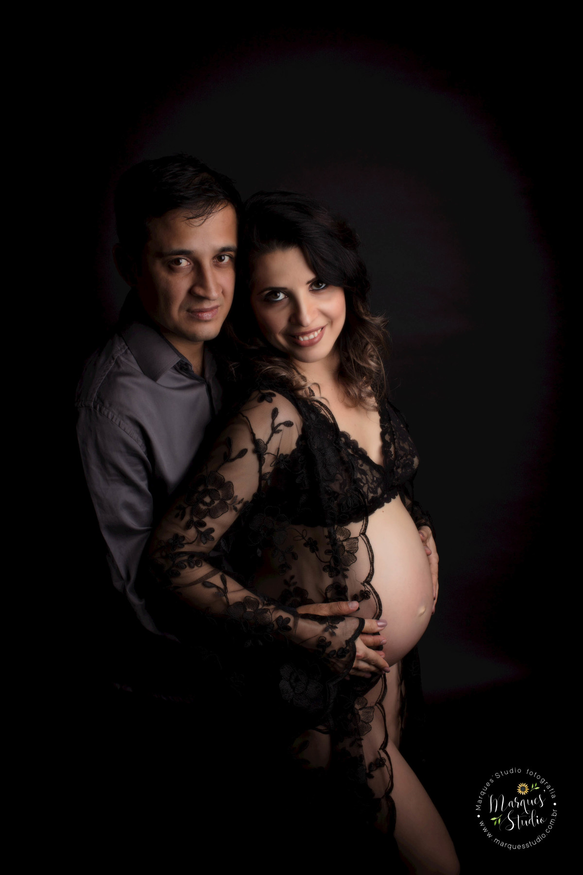 Fotografia de studio fotográfico feita no Sumaré São Paulo SP com um casal de grávidos eles estão abraçados ela está na sua frente e ambos estão com as mãos sobre a barriga da gestante com um lindo sorriso ela usa uma camisola preta ele usa calça e camisa