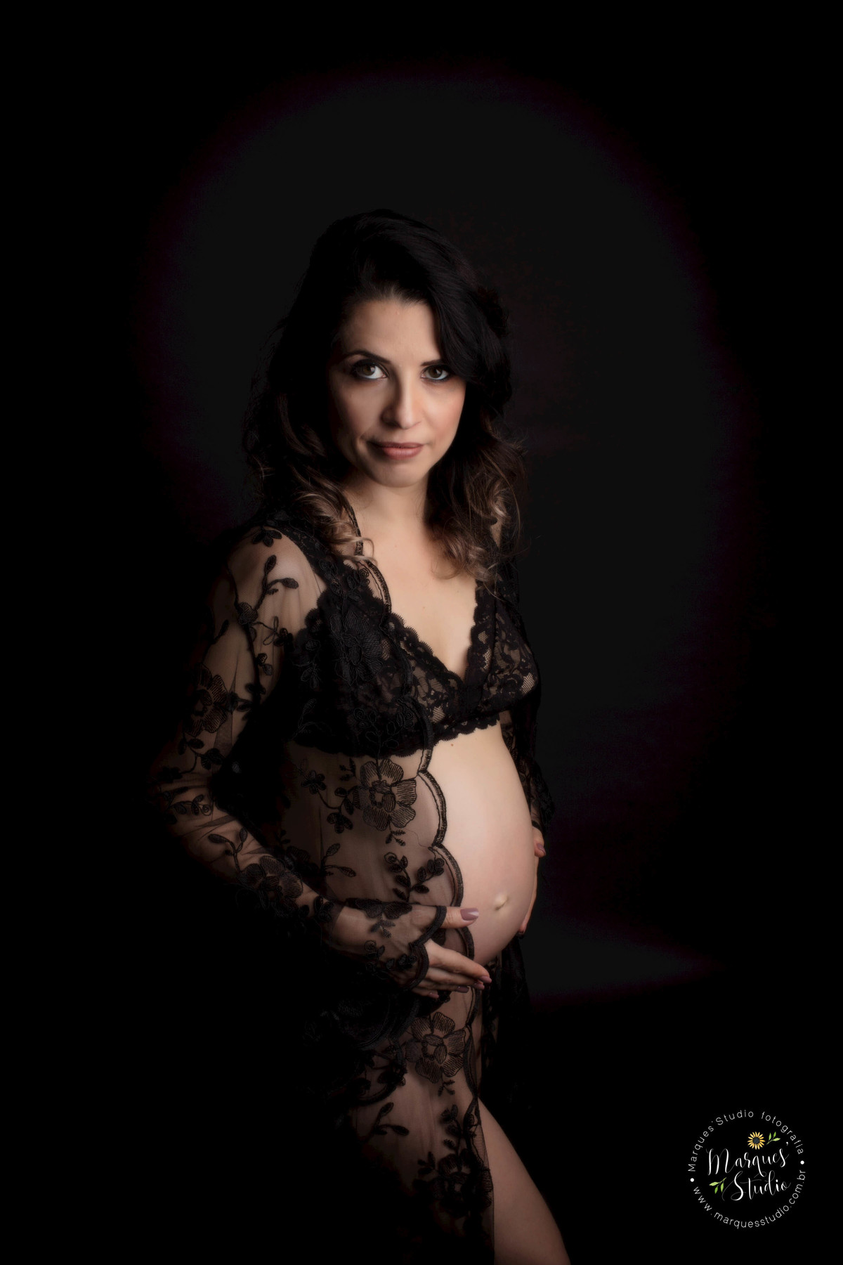 Fotografia em Studio da gestante Camila, localizado na Avenida Paulista, São Paulo SP, ela está de perfil e em pé, com as mãos em sua barriga de gravida, no fundo infinito preto, ela está usando uma linda camisola preta de renda.