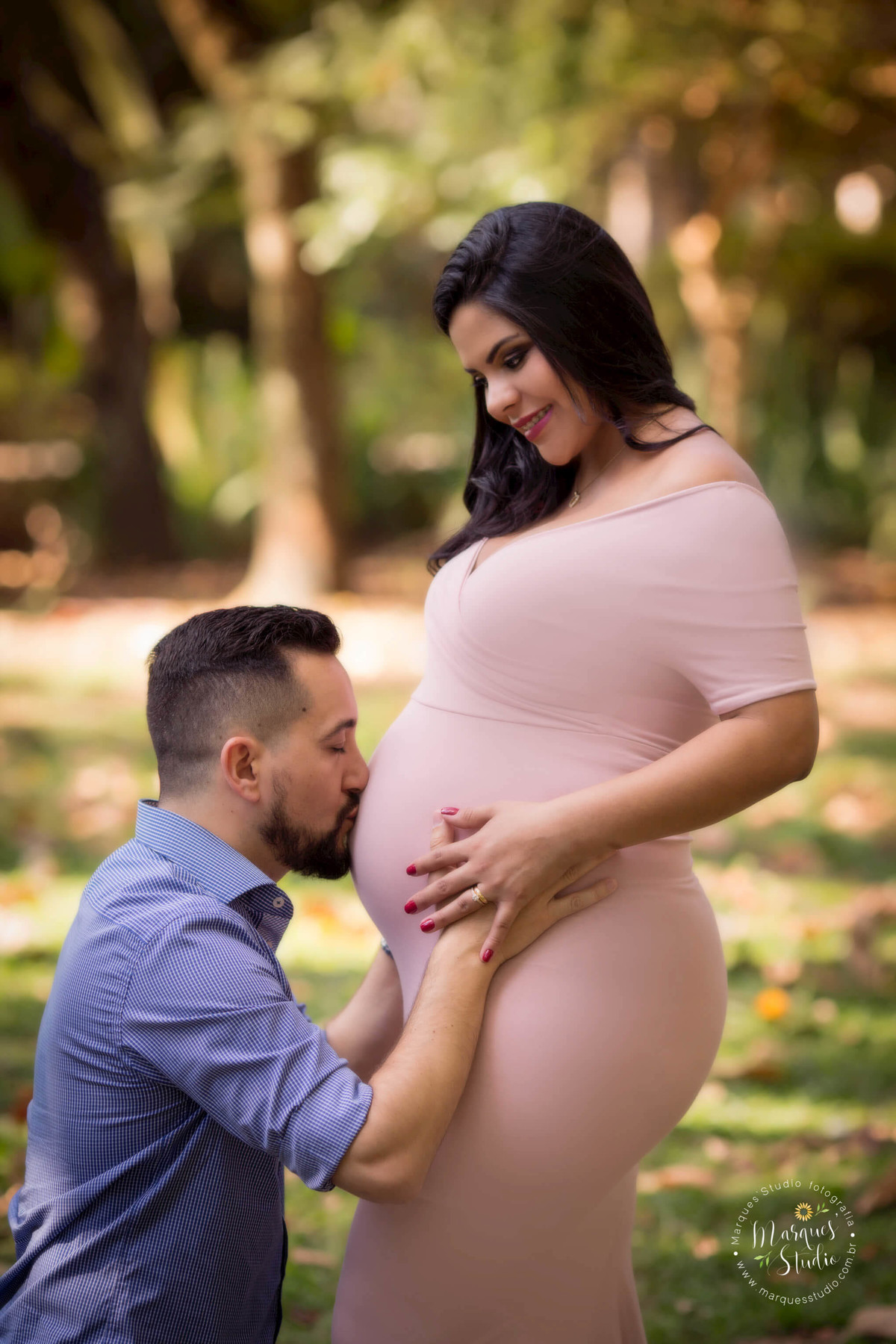 Fotografia do ensaio gestante da Thamyres, na foto ela está de frente para seu esposo, ele está dando um beijo na sua barriga de gestante, eles estão no parque na Avenida Paulista, São Paulo, SP, ela sorri para ele, ao fundo tem uma linda arvore.
