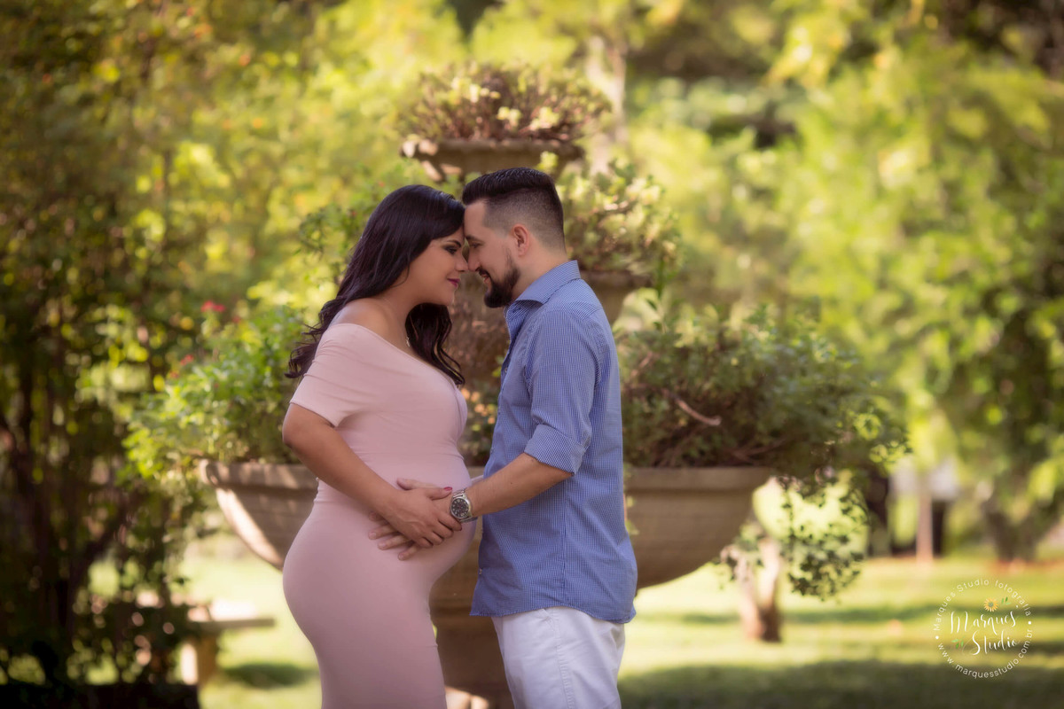 Fotografia de ensaio gestante externa, no parque Burle Marx, São Paulo, SP, a gestante está com o seu esposo um de frente para o outro bem abraçados, ao fundo uma linda área verde com muitas flores e arvores, ela usa um vestido rosa e ele calça e camisa.