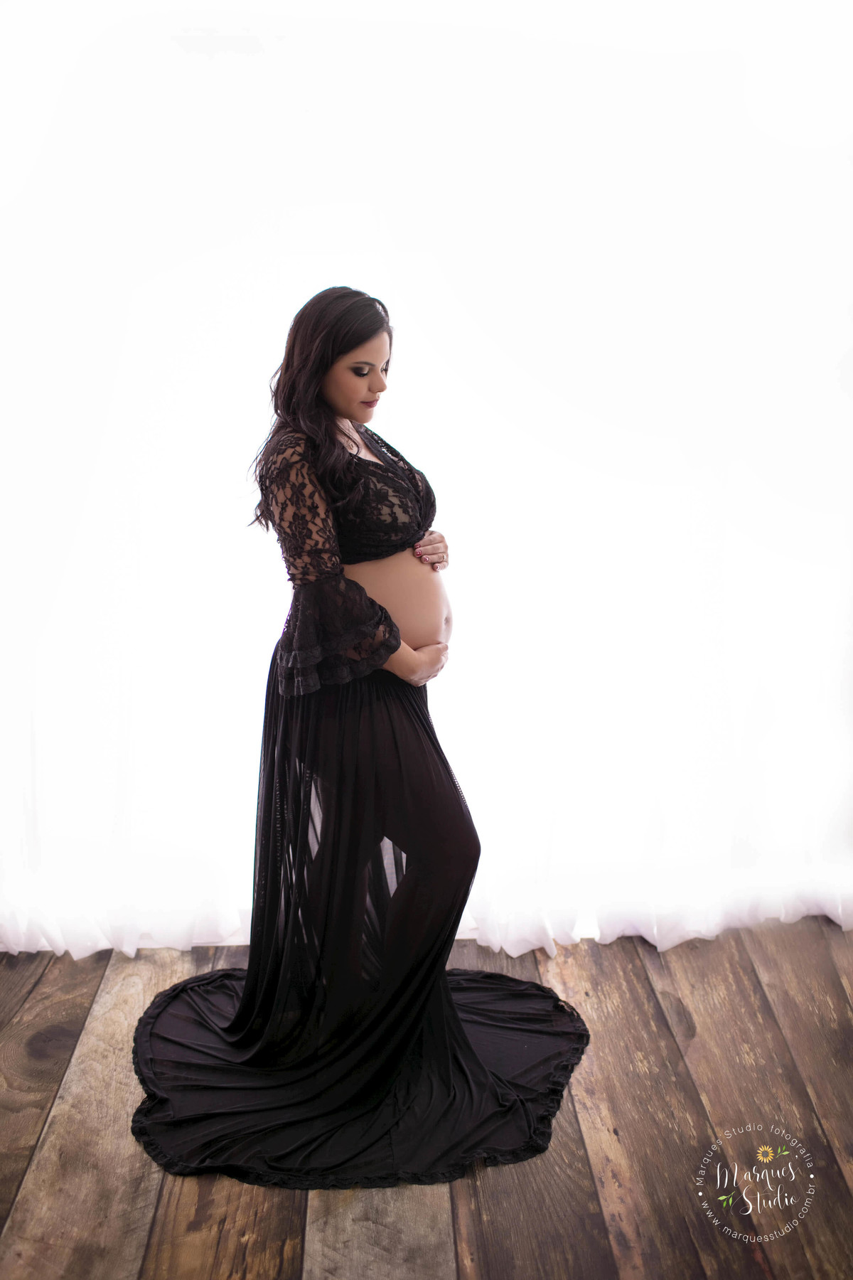 Gestante linda em seu ensaio fotográfico no studio localizado em Osasco, São Paulo, SP, com o olhar para o seu bebê, ela está com as duas mãos sobre sua barriga em um lindo fundo fotográfico branco, com madeira, ela usa um conjunto de top e saia preto.