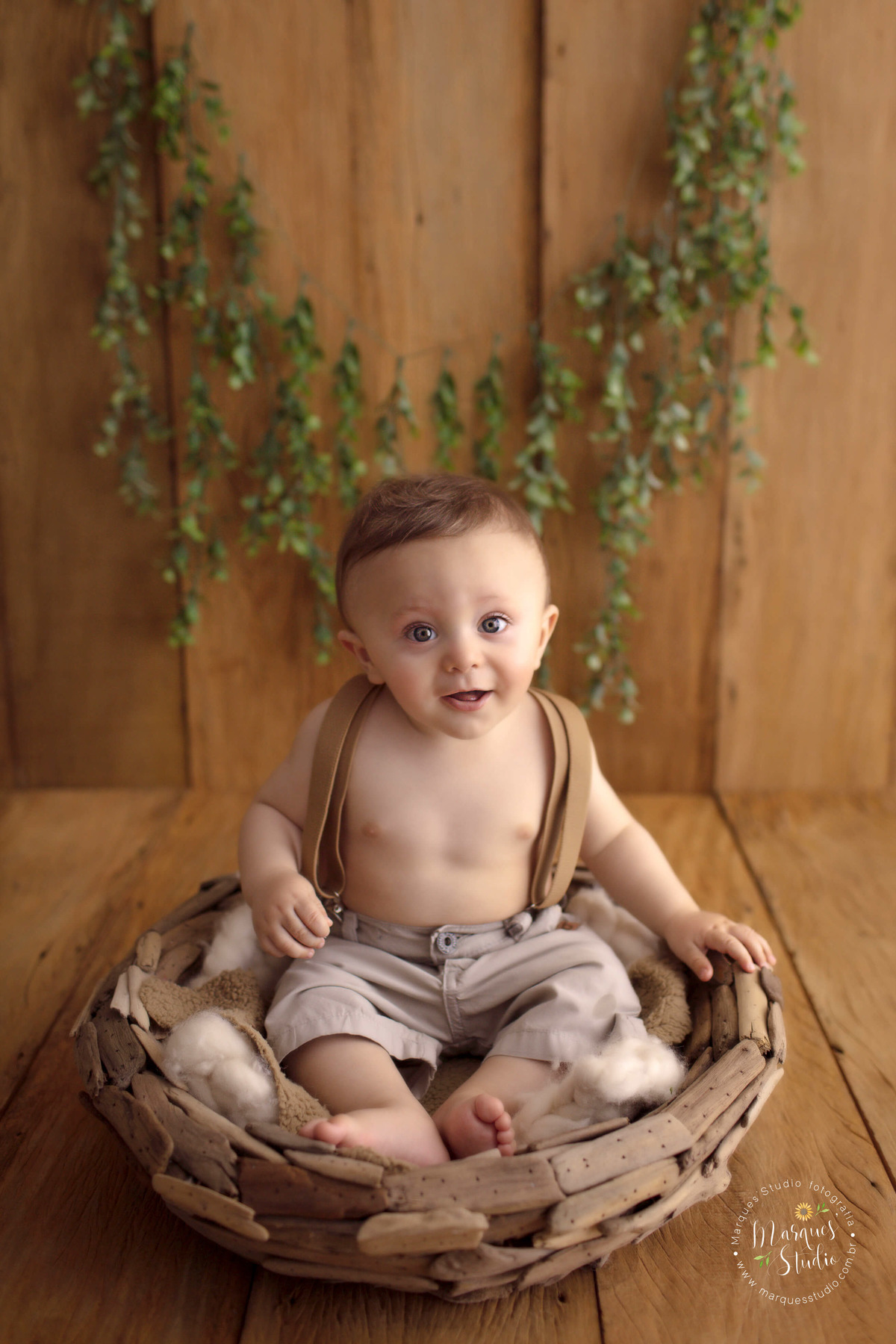 Foto Feita para a sessão de acompanhamento infantil em studio, no Morumbi, São Paulo, SP, de um bebê de 8 meses sorrindo, ele está sentado em um cesto de madeira, e o fundo fotográfico é de madeira com folhas verdes, ele está de short e suspensório bege.