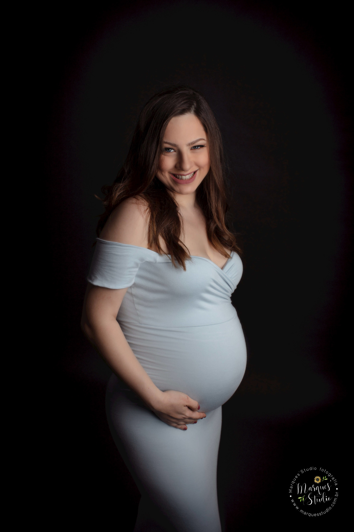 Foto de Studio da gestante Patricia, localizado na Avenida Paulista, São Paulo SP, ela está de perfil e em pé, com as mãos em sua barriga de gravida, no fundo infinito preto, com um lindo sorriso no rosto, ela usa um lindo vestido longo na cor azul claro.