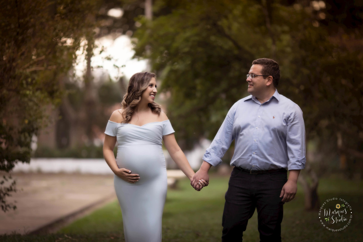 Foto de um lindo casal de grávidos no parque, eles estão de mãos dadas, se olhando e sorrindo, ela está usando um lindo vestido longo azul claro, no nosso studio na Vila Leopoldina, SP, temos vários modelos e cores de vestidos disponíveis para os ensaios.