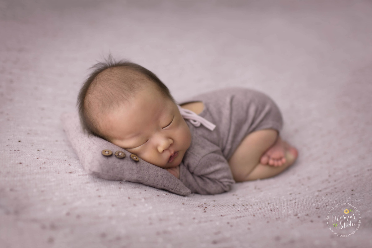Fotografia de bebê recém-nascido menino, deitado no puff com as mãozinhas no rosto, foto feita em studio fotográfico na Vila Perdizes, São Paulo SP, ele está dormindo e usando uma roupinha marrom, com o rosto em um lindo travesseiro marrom, Newborn gêmeos