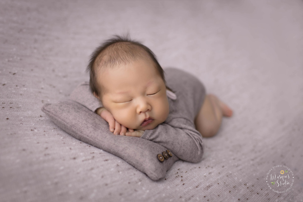 Fotografia de bebê menino, deitado no puff com as mãos no rosto, foto feita em studio fotográfico na Avenida Paulista, São Paulo, SP, ele está dormindo e usando um lindo macacão na cor marrom, e está sobre um lindo travesseiro marrom, Newborn de gêmeos.