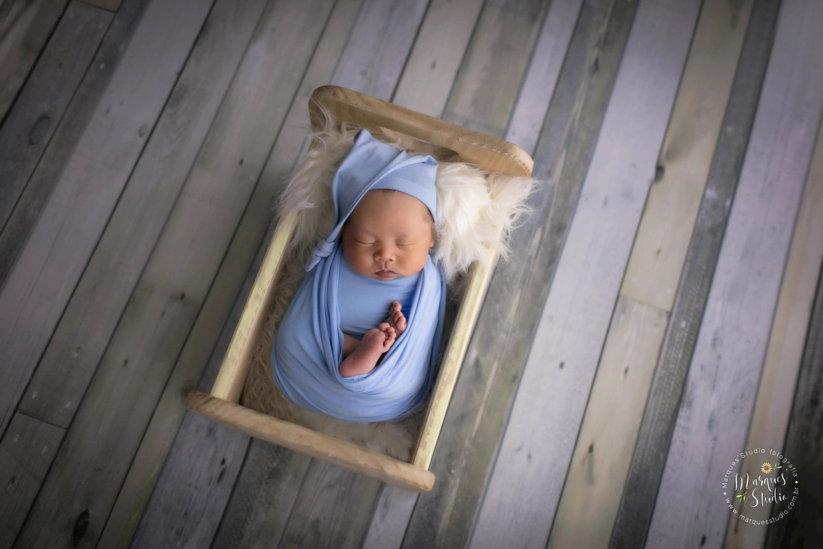 Ensaio newborn feito no nosso studio na Vila Leopoldina, São Paulo, SP. Bebê no wrap, de barriga pra cima dentro do berço. No fundo fotográfico de madeira. Ele usa wrap e touca azul claro, foto de recém-nascidos gêmeos.