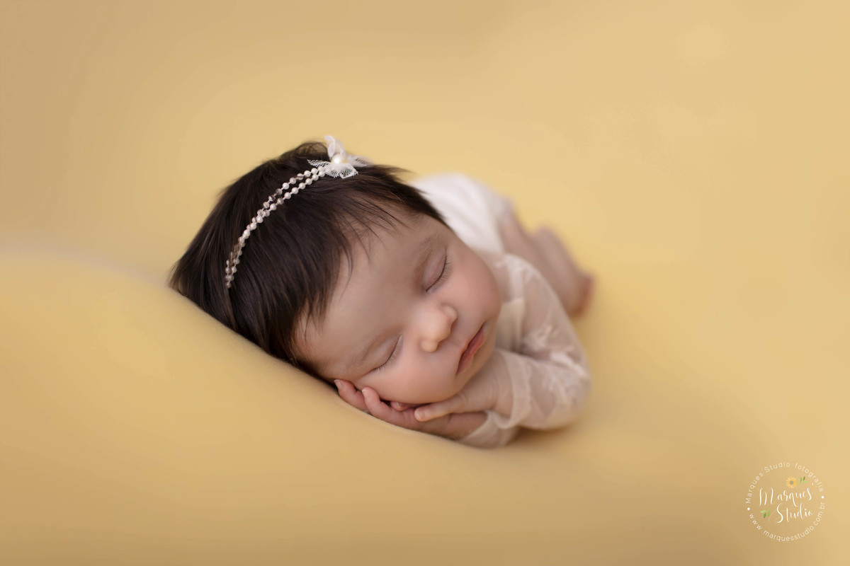 Foto de bebê menina, deitada no puff com uma manta amarela, com as mãos no rosto, foto feita em studio fotográfico localizado na Vila Leopoldina, São Paulo, SP, ela está dormindo e usando um lindo macacão na cor branca com renda e lacinho de pérolas.