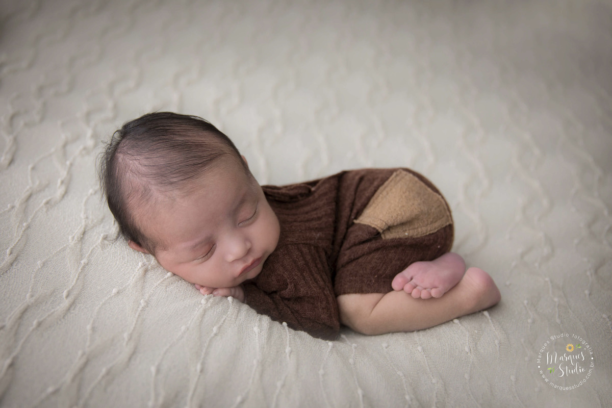Foto de bebê menino, deitado no puff com uma manta bege, com as mãos no rosto, foto feita em studio fotográfico localizado na Vila Leopoldina, São Paulo, SP, ele está dormindo e usando um lindo macacão na cor marrom com bolso bege.