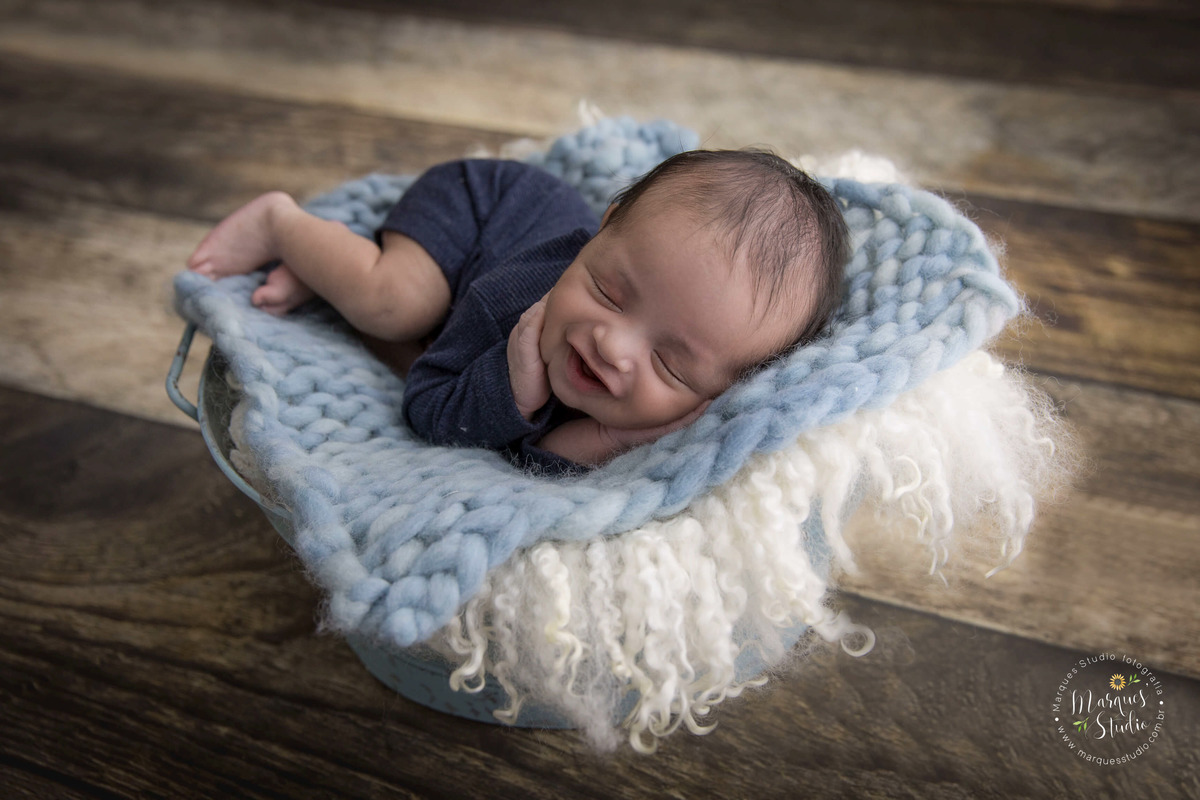Foto feita para o ensaio Newborn do Davi, feito no nosso studio localizado na Vila Sumaré, São Paulo, SP. O Bebê está deitadinho em um baldinho com mantas azuis, no fundo fotográfico de madeira, ele está dando um lindo sorriso enquanto dorme.