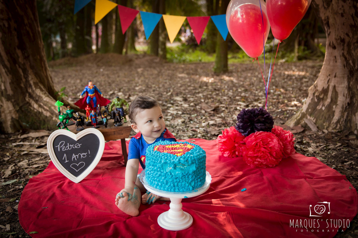 Fotografia de Bebê de 12 meses no ensaio Smash The Cake no parque localizado em São Paulo SP com um lindo bolo azul personalizado do super homem, balões vermelhos, e bandeiras coloridas ao fundo, o bebê está sentado sorrindo ao lado dos personagens Marvel