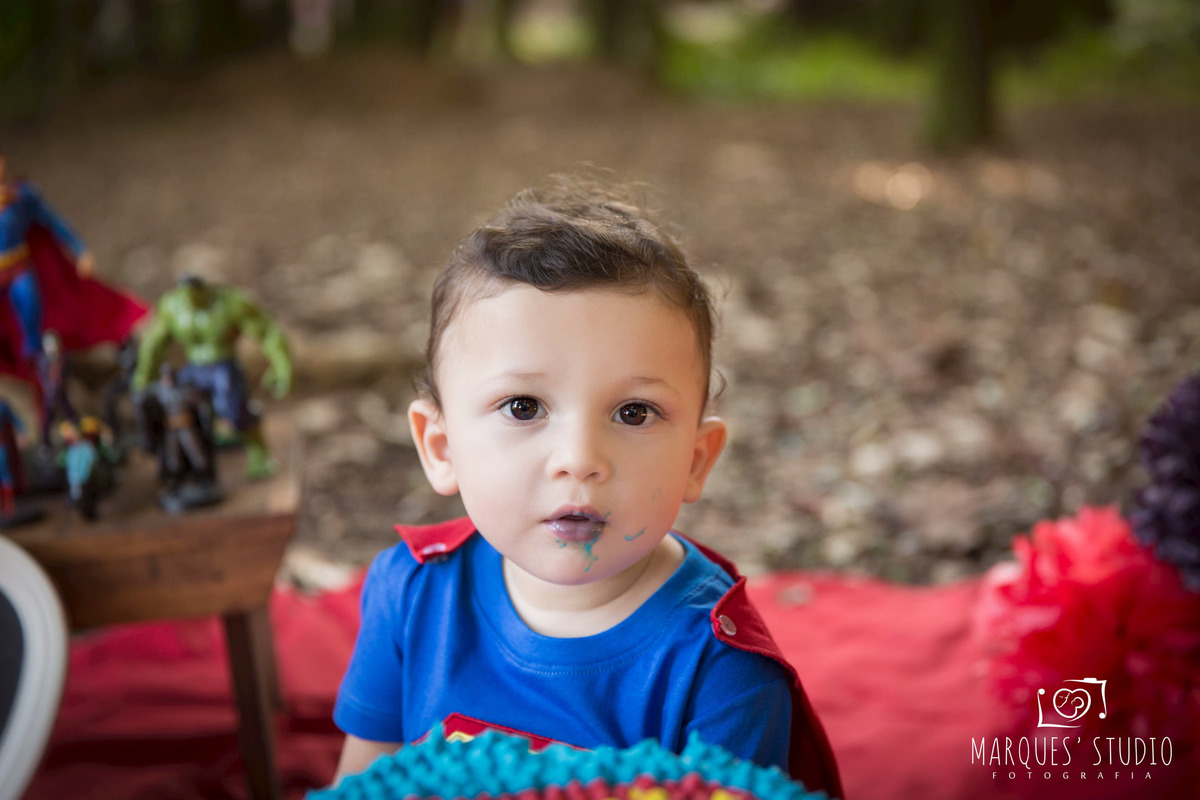 Fotografia para a sessão Smash The Cake externa com o tema do super homem, na Vila Sumaré, São Paulo, SP, de uma criança de 12 meses sorrindo sentado na grama de um parque, com balões e bandeirinhas ao fundo, ele usa a roupa personalizada do super homem.