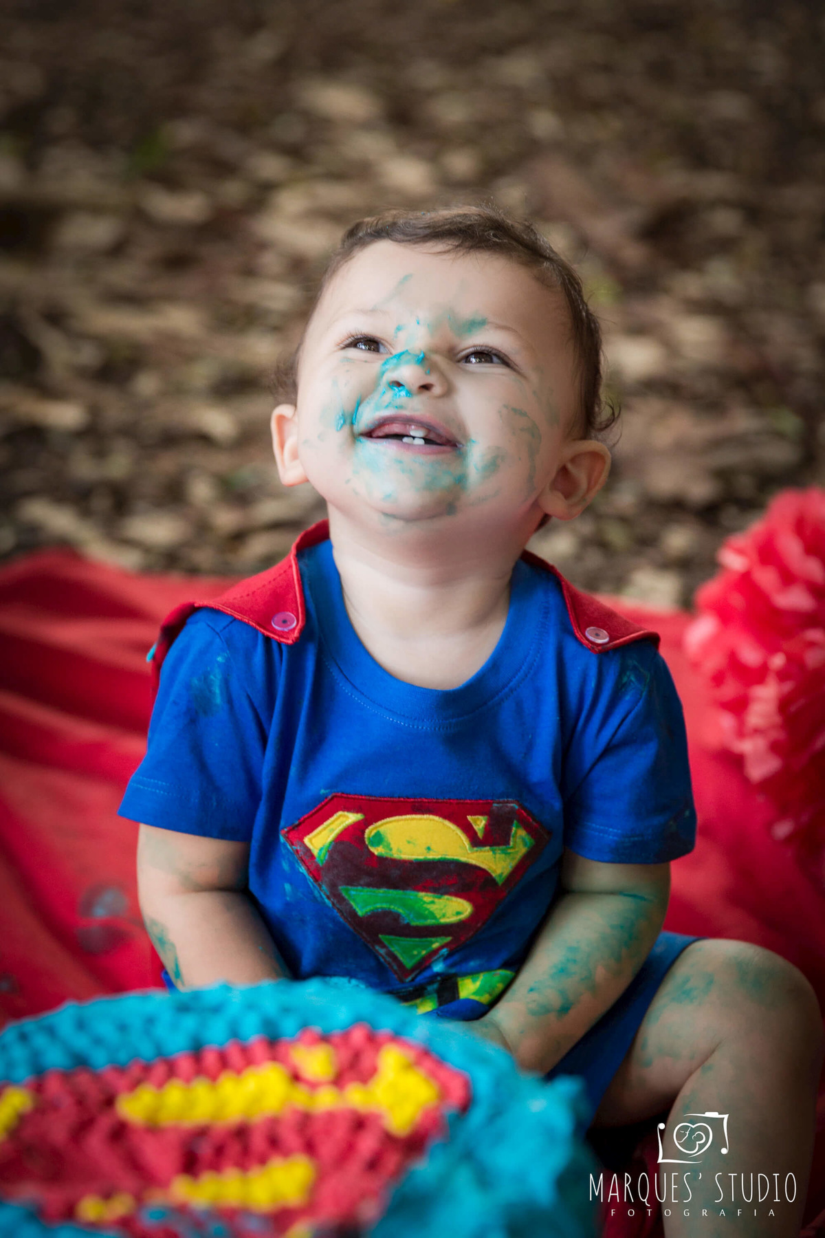 Foto Feita para a sessão Smash The Cake externa com o tema do super homem, na Vila Leopoldina São Paulo, SP, de uma criança de 1 ano sorrindo sentado na grama de um parque, com balões e bandeirinhas ao fundo, ele usa a roupa personalizada do super homem.