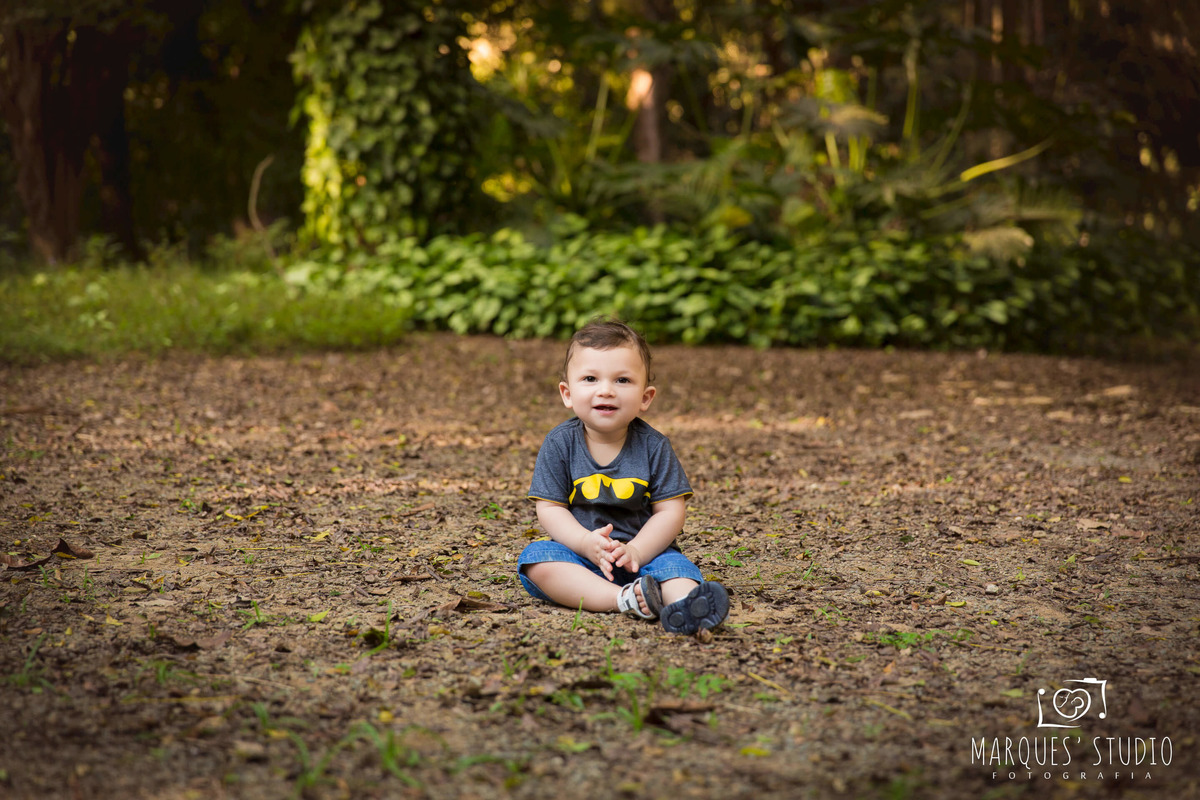 Ensaio feito para o acompanhamento de 12 meses do Pedro, ele está em um belíssimo parque na Vila Leopoldina, São Paulo, SP, ele está sentado sobre a grama e as folhas secas, dando um sorriso, ele está usando uma linda camiseta do personagem Batman e short