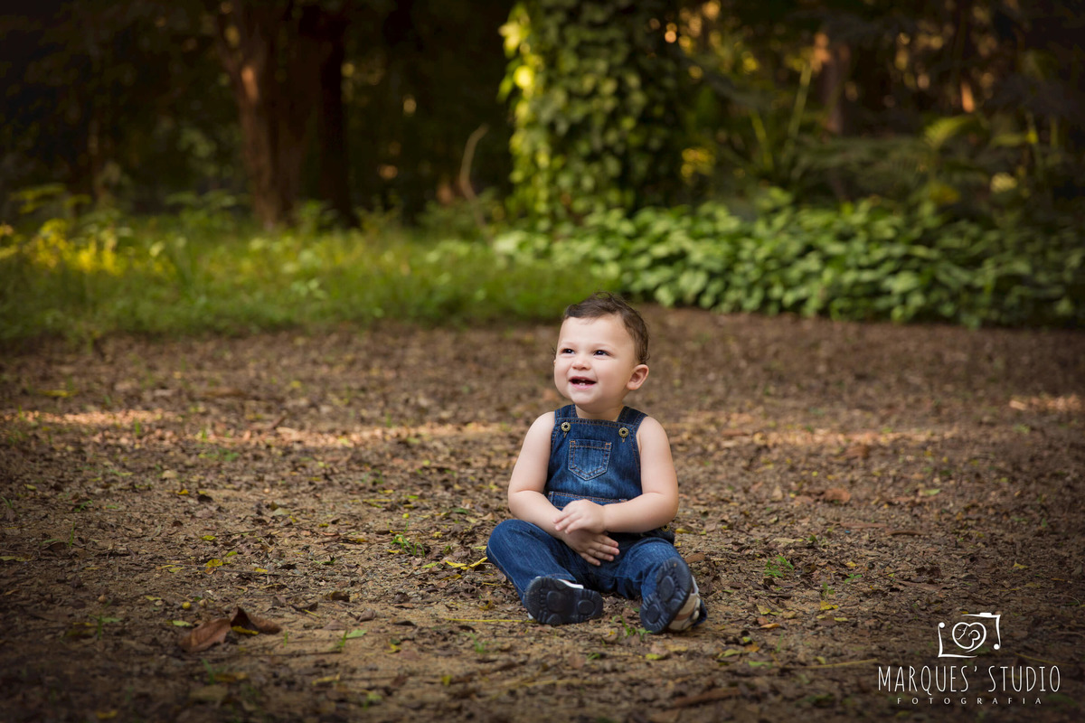 Foto feita para o acompanhamento de 12 meses do Pedro, ele está em um belíssimo parque na Vila Madalena, São Paulo, SP, ele está sentado sobre a grama e as folhas secas, dando um lindo sorriso, ele está usando um lindo macacão jeans.