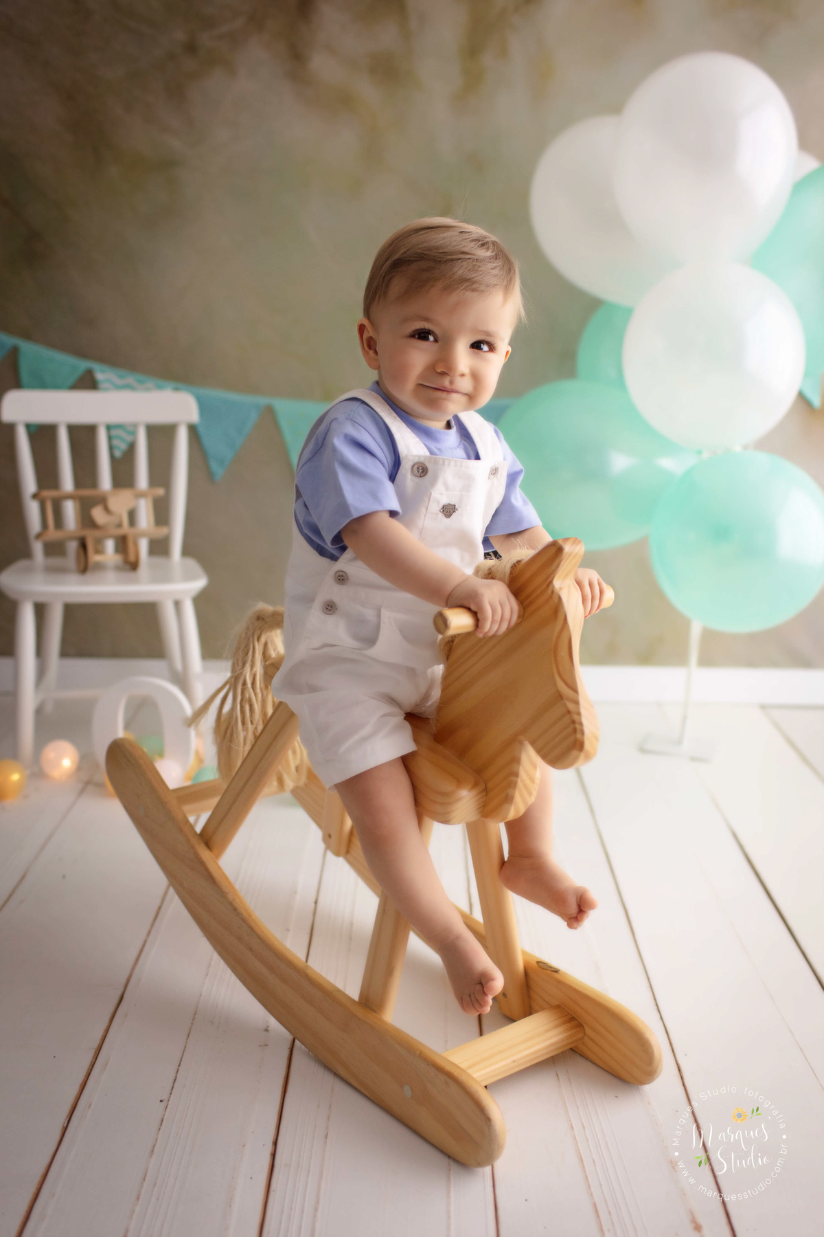 Foto de um bebê de 12 meses na sua sessão de acompanhamento, ele está sentado em um lindo cavalinho de madeira, ao fundo temos uma cadeirinha branca com um avião, bandeiras e balões verde e branco, o studio fica localizado na Vila Leopoldina São Paulo SP.