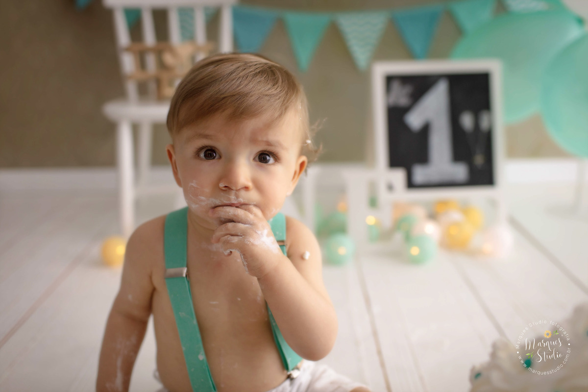 Foto Feita para a sessão Smash The Cake do Leo, no Studio na Av. Paulista SP, ele está com 1 aninho, está sentado e com sua mão esquerda na boca, com balões e bandeirinhas ao fundo, ele está com um conjunto de short branco e suspensório verde Tiffany
