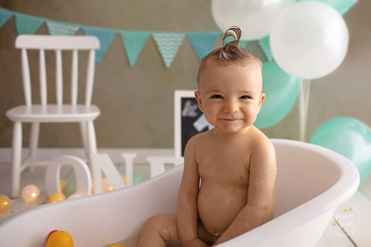 Foto do bebê Leo de 12 meses na sua sessão de acompanhamento, ele está sentado em uma linda banheira branca, tomando banho e está sorrindo, ao fundo tem uma cadeirinha branca a palavra one, bandeiras e balões verde e branco, o studio fica em Osasco, SP.