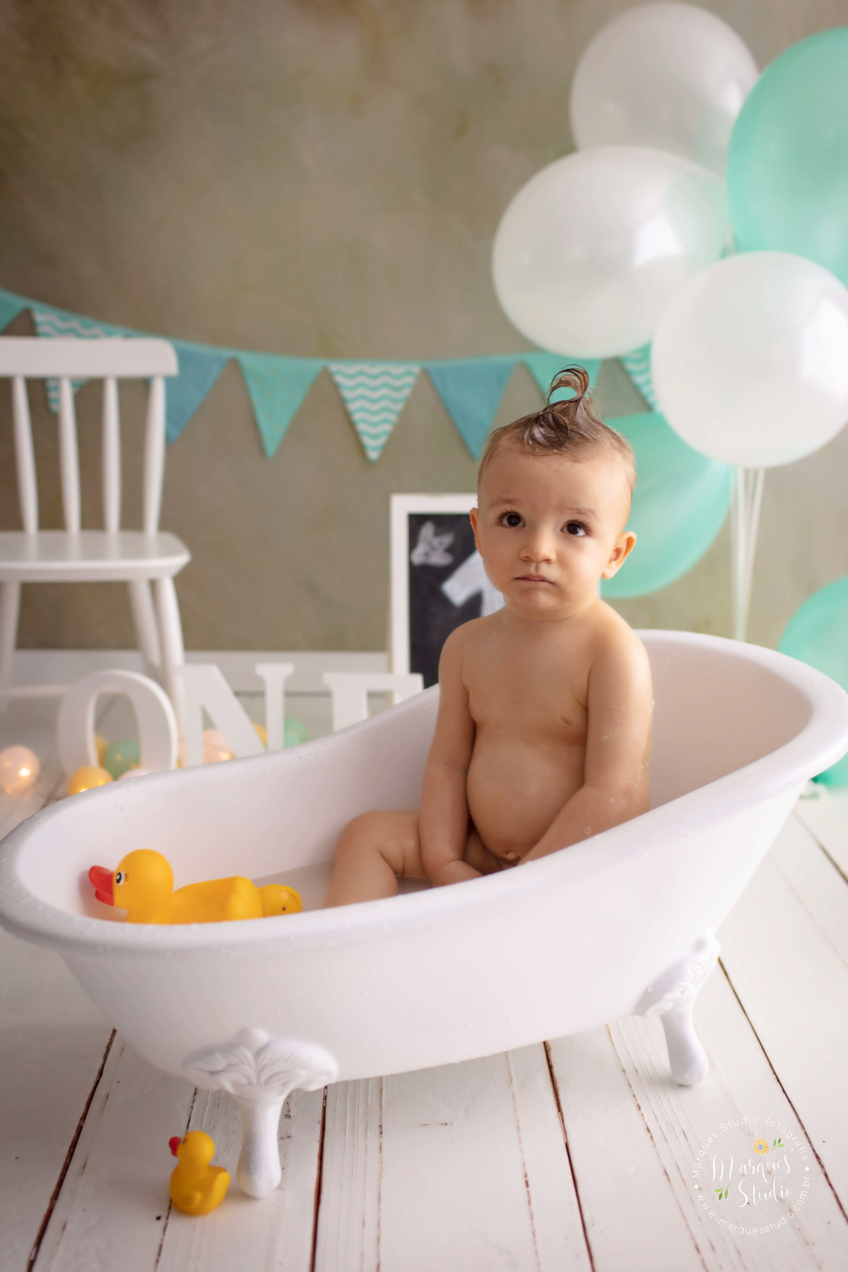 Fotografia do Leo de 12 meses na sua sessão de acompanhamento, ele está sentado em uma linda banheira branca, tomando banho, ao fundo tem uma cadeirinha branca a palavra one, bandeiras e balões verde e branco, o studio fica na Vila Leopoldina, SP.
