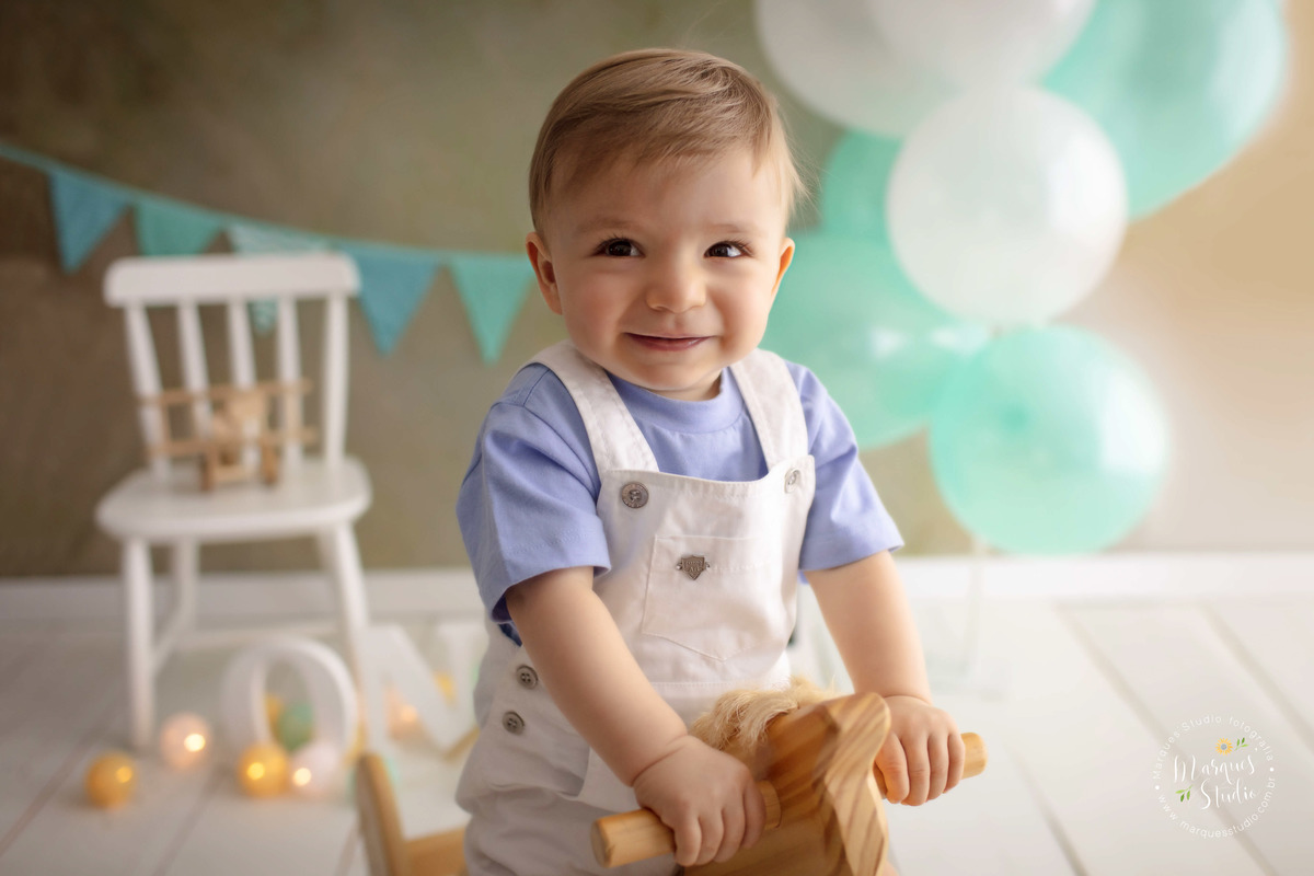 Foto do bebê Leo de 12 meses na sua sessão de acompanhamento, ele está sentado em um lindo cavalinho de madeira, e está sorrindo, ao fundo tem uma cadeirinha branca com um avião, bandeiras e balões verde e branco, o studio fica na Capital São Paulo SP.