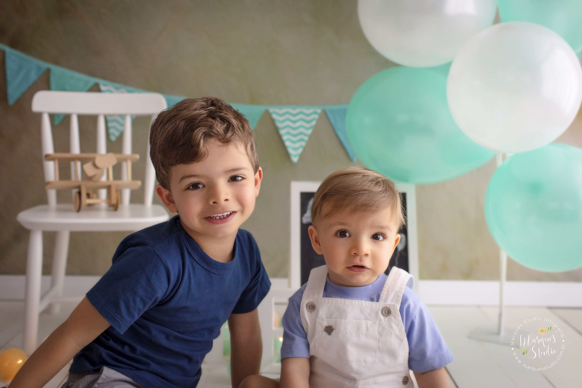 Fotografia de dois irmãos sentados em um fundo fotográfico de madeira, eles estão com um lindo sorriso no rosto, o ensaio foi feito no estúdio na Vila Leopoldina, São Paulo, SP, para a sessão de acompanhamento do Leo, ao fundo tem bandeiras e balões. 