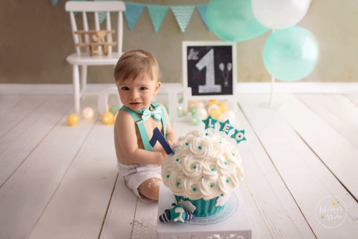 Fotografia do Bebê Leo de 12 meses no ensaio Smash The Cake no studio localizado na Vila Sumaré São Paulo SP ele está atras do bolo nas cores verde e branco com um aviãozinho decorativo, ao fundo uma cadeira branca balões e bandeiras verdes e branco.