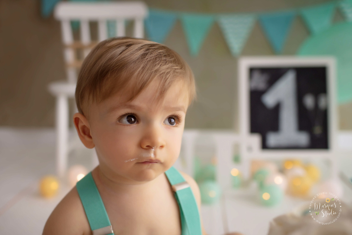 Foto Feita para a sessão Smash The Cake no studio, na zona Oeste, Osasco, SP, de uma criança de 1 ano ele está sentado e olhando para a esquerda dele, com balões e bandeirinhas ao fundo, ele está com um conjunto de short branco, e suspensório verde.