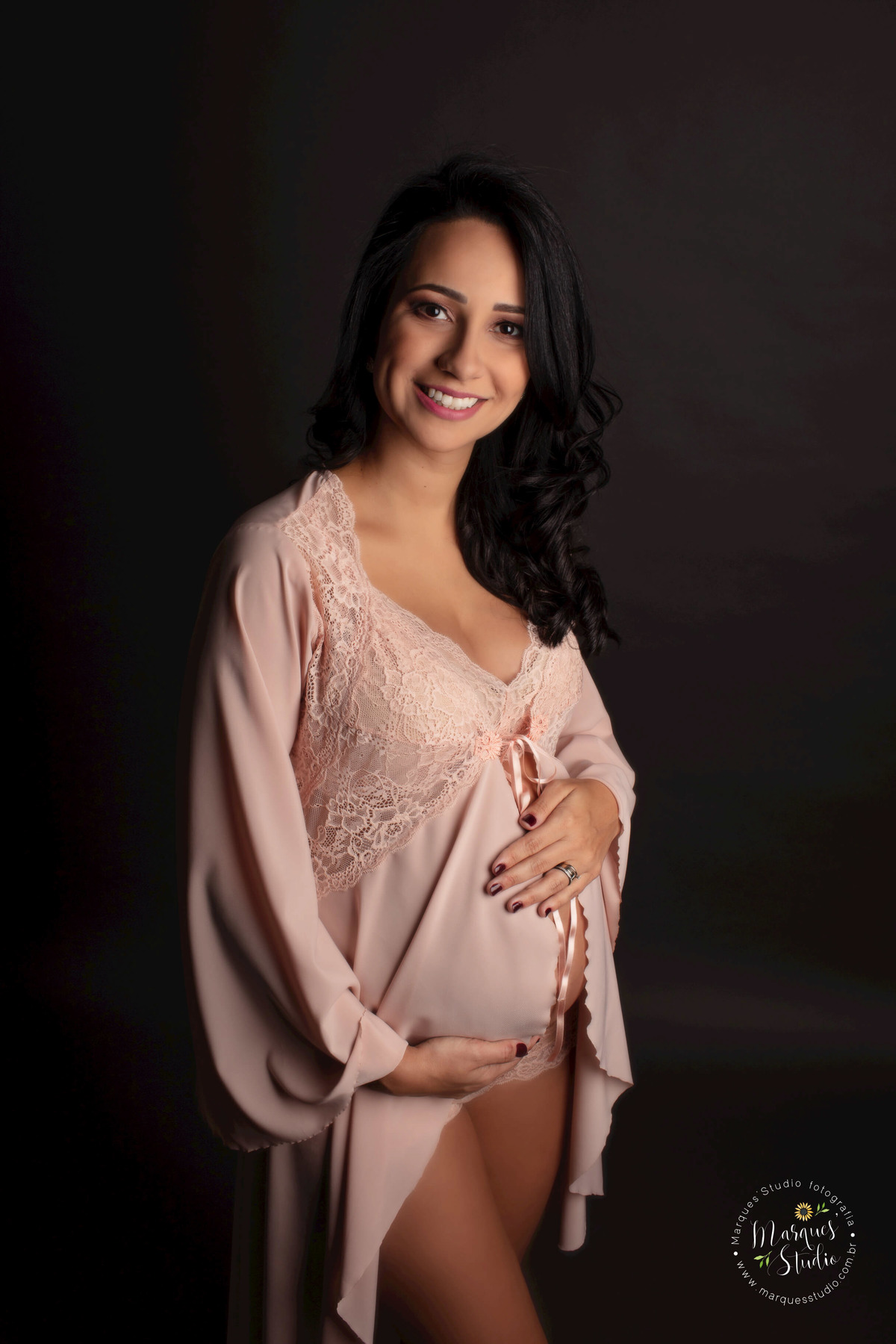 Foto de Studio da gestante Paula, localizado na Avenida Paulista, São Paulo SP, ela está de perfil e em pé, com as mãos em sua barriga de gravida, no fundo infinito preto, com um lindo sorriso no rosto, ela usa uma linda camisola na cor rose com renda.
