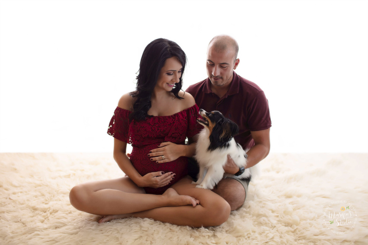 Foto de um casal de grávidos com sua cachorrinha para o ensaio de gestante da Paula, eles estão se olhando e felizes todos estão sentados em um tapete bege, a mamãe usa um lindo body vermelho e o papai short e camisa polo vermelha, estão no estúdio  em SP