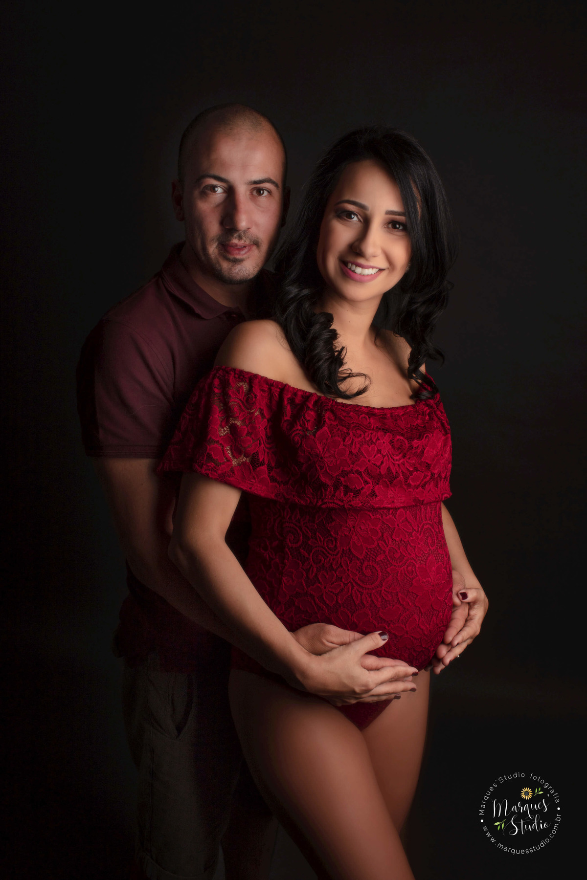 Fotografia de um casal de grávidos, a mamãe está na frente do papai, os dois estão com as mãos sobre a barriga da gestante, eles estão sorrindo, ela usa um body vermelho, e ele usa camiseta e short jeans, eles estão no studio em Pinheiros São Paulo, SP.