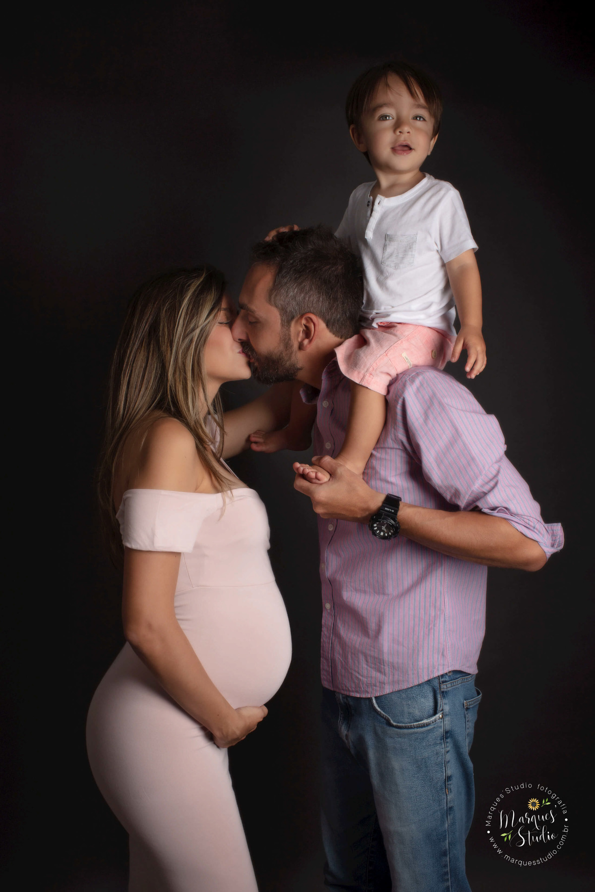 Foto de uma família que estão no estúdio na Av. Paulista, São Paulo, SP, a mamãe está dando um beijo no papai, e seu filho mais velho está nas costas do papai, essa foto foi feita para o ensaio gestante da Camila.