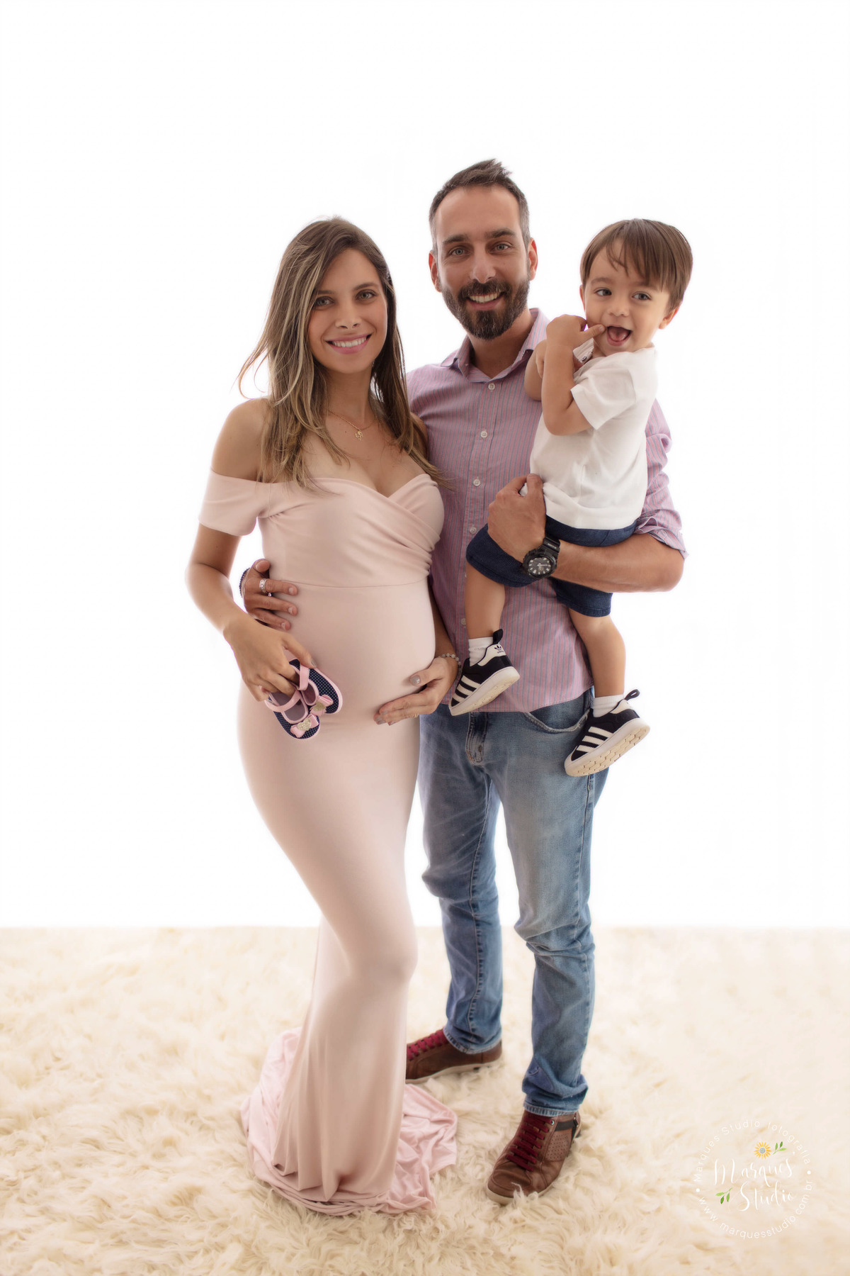 Fotografia de um casal de grávidos e seu bebê, a mamãe está ao lado do papai, o pai está abraçando a mãe e com o irmão mais velho no colo, eles estão sorrindo, foto feita no studio localizado na Av. Paulista, São Paulo, SP.