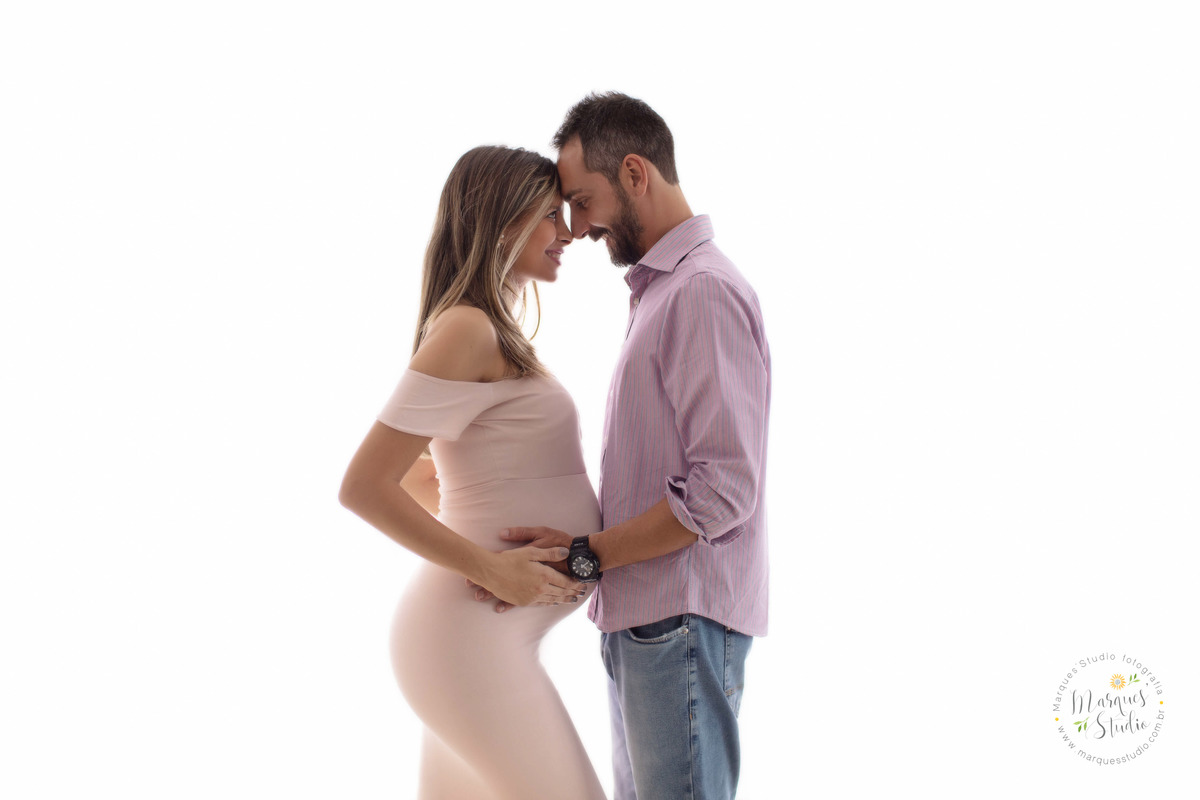 Fotografia feita no studio na Capital São Paulo SP com um lindo casal de grávidos eles estão um de frente para o outro colocando as mãos sobre a barriga da gestante ela usa um lindo vestido longo rosa, ele calça e camisa, e estão no fundo infinito branco.