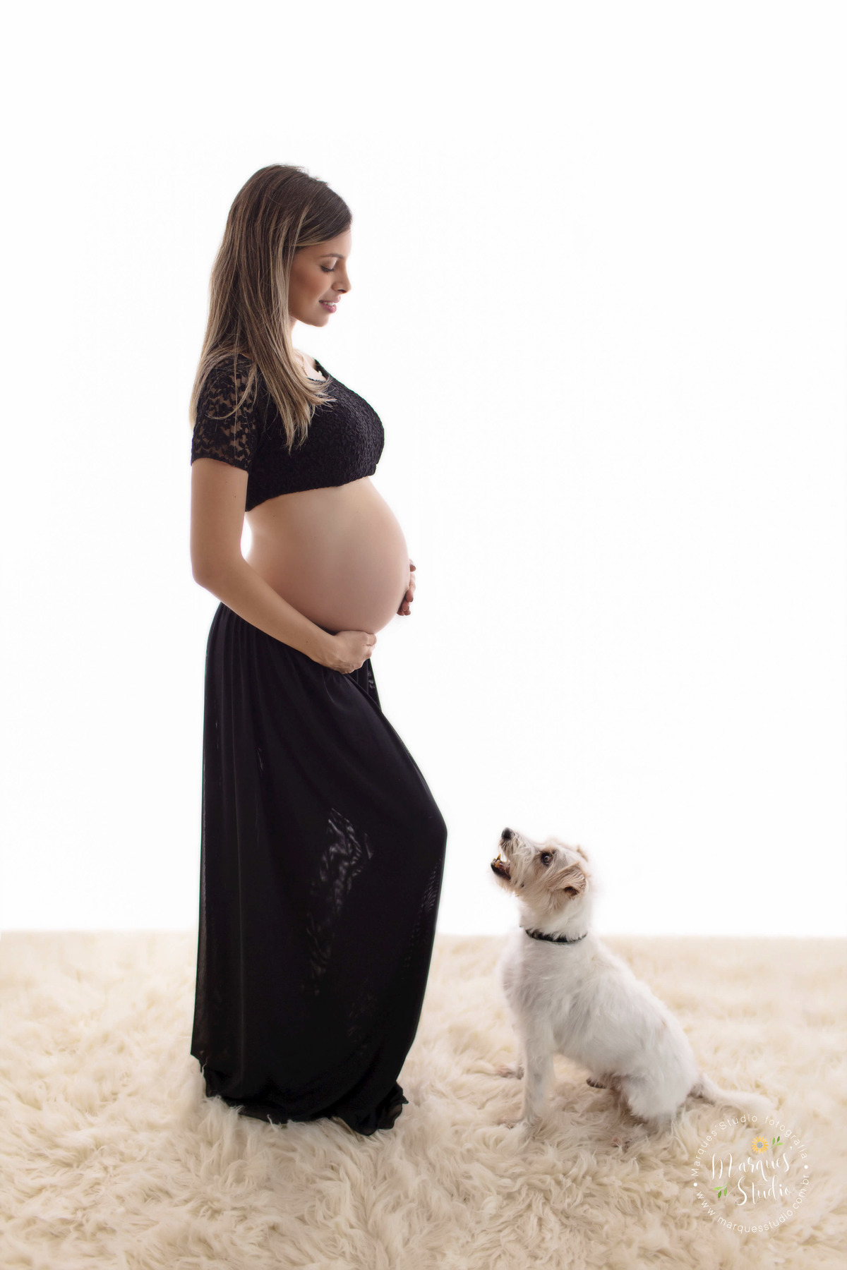 Foto feita em studio fotográfico localizado na Vila Madalena São Paulo SP para o ensaio gestante da Camila, ela está usando um lindo conjunto de top e saia preto, ela está de perfil com as mãos sobre sua barriga, de frente para ela está o seu cachorrinho.