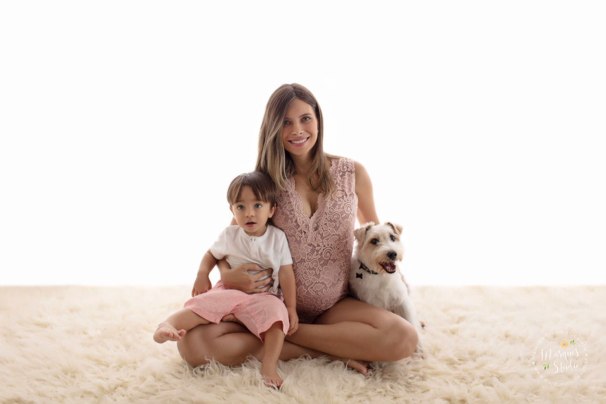 Foto feita no ensaio da gestante Camila, no studio localizado na Pompéia, São Paulo, SP, na foto a gestante está sentada no tapete bege, e em seu colo está seu filho mais velho e seu cachorrinho lindo, eles estão felizes e sorridentes.